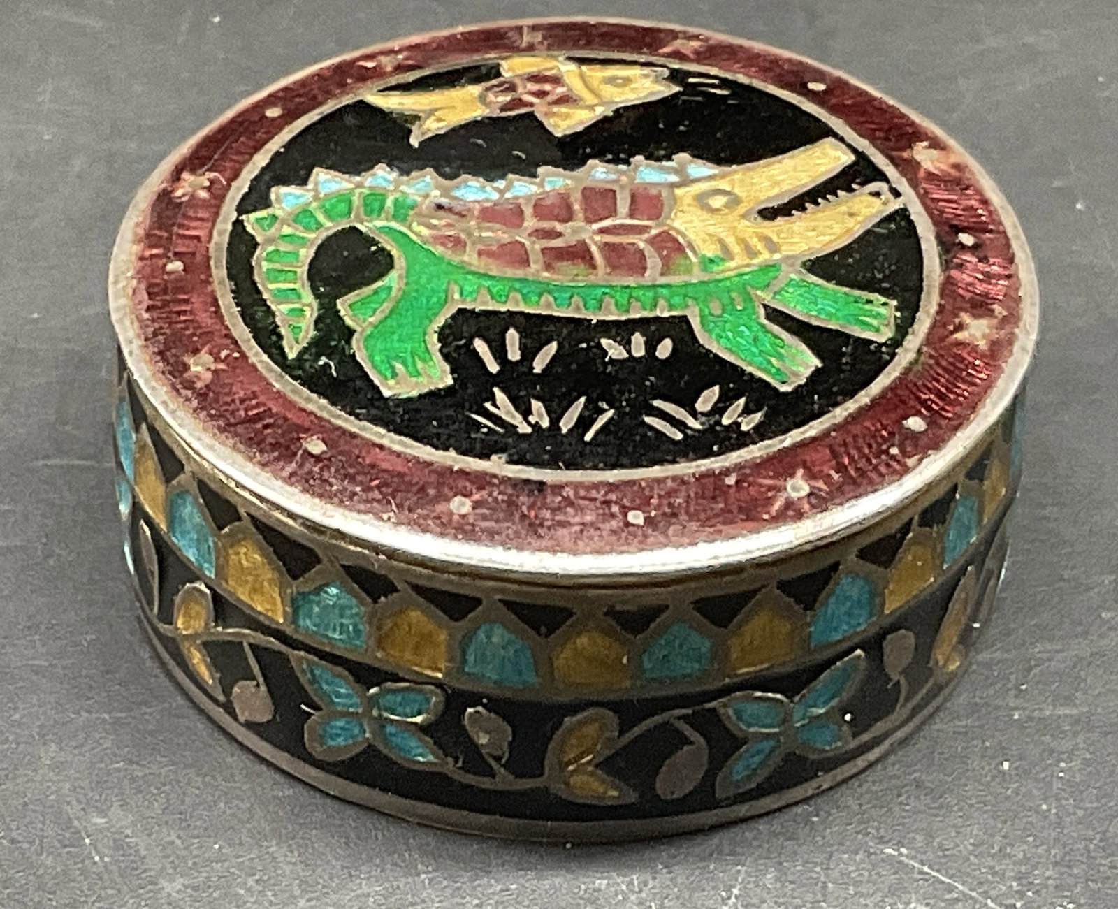 VTG Mexican Cloisonne Enamel Alligator Pill Box: Vintage Circa 1970s-1990s Mexican Cloisonne Enamel Alpaca Silver Pill Box. Colorful Alligator and Fish Folk Art Design. Blue and Yellow Floral Design on Edges. Comes in Silk Case. Scratching and Tarni