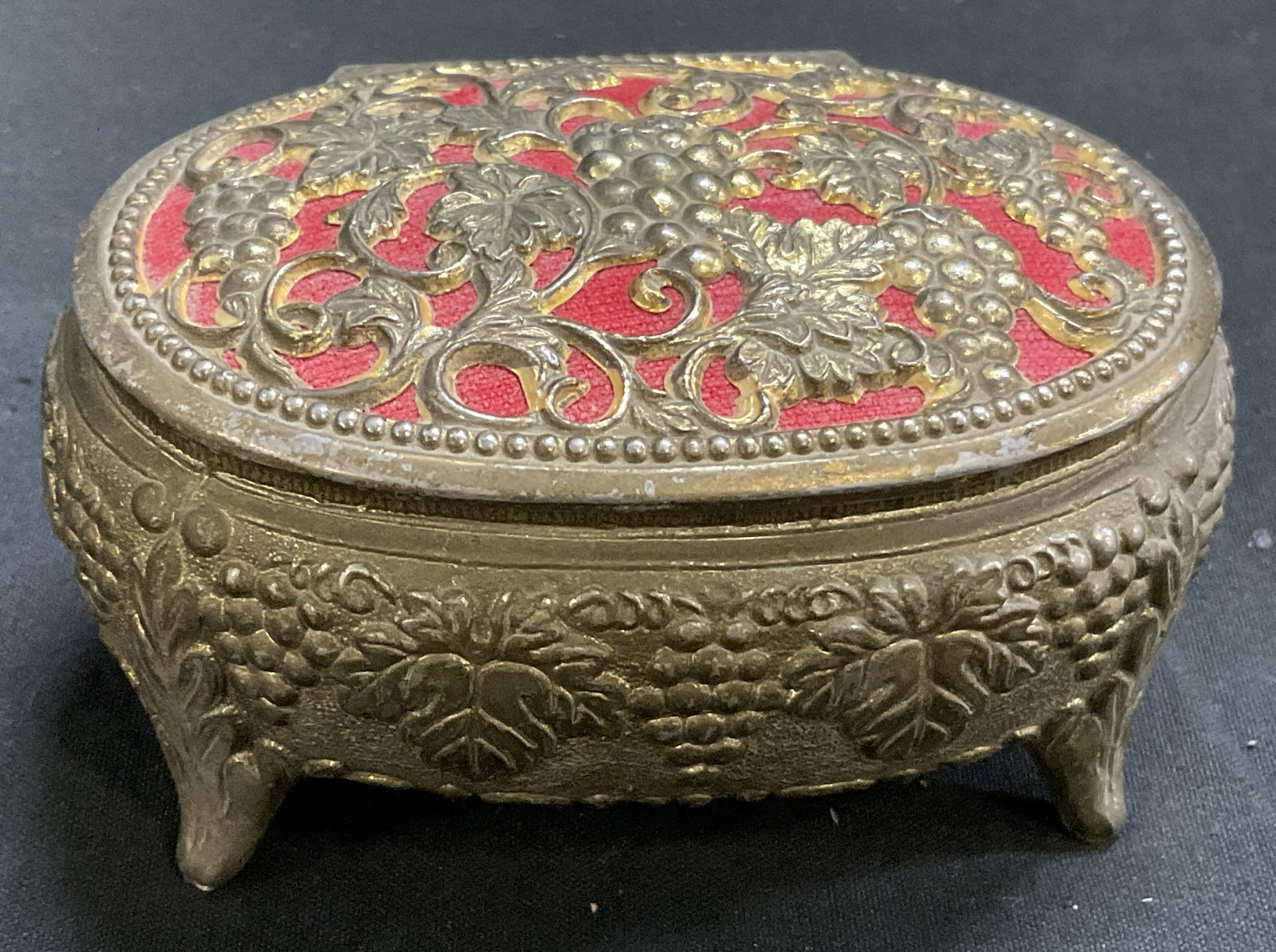 VTG Brass-Toned Filigree Footed Trinket Box JPN: Vintage Brass-Toned Footed Trinket Box. Grape and Vine Leaf Filigree Design. Red Fabric Backdrop. Red Velvet Lining. Japan. Some Losses to Coating and Aging to Metal. Measures app 1.75in H x 3in Diam.