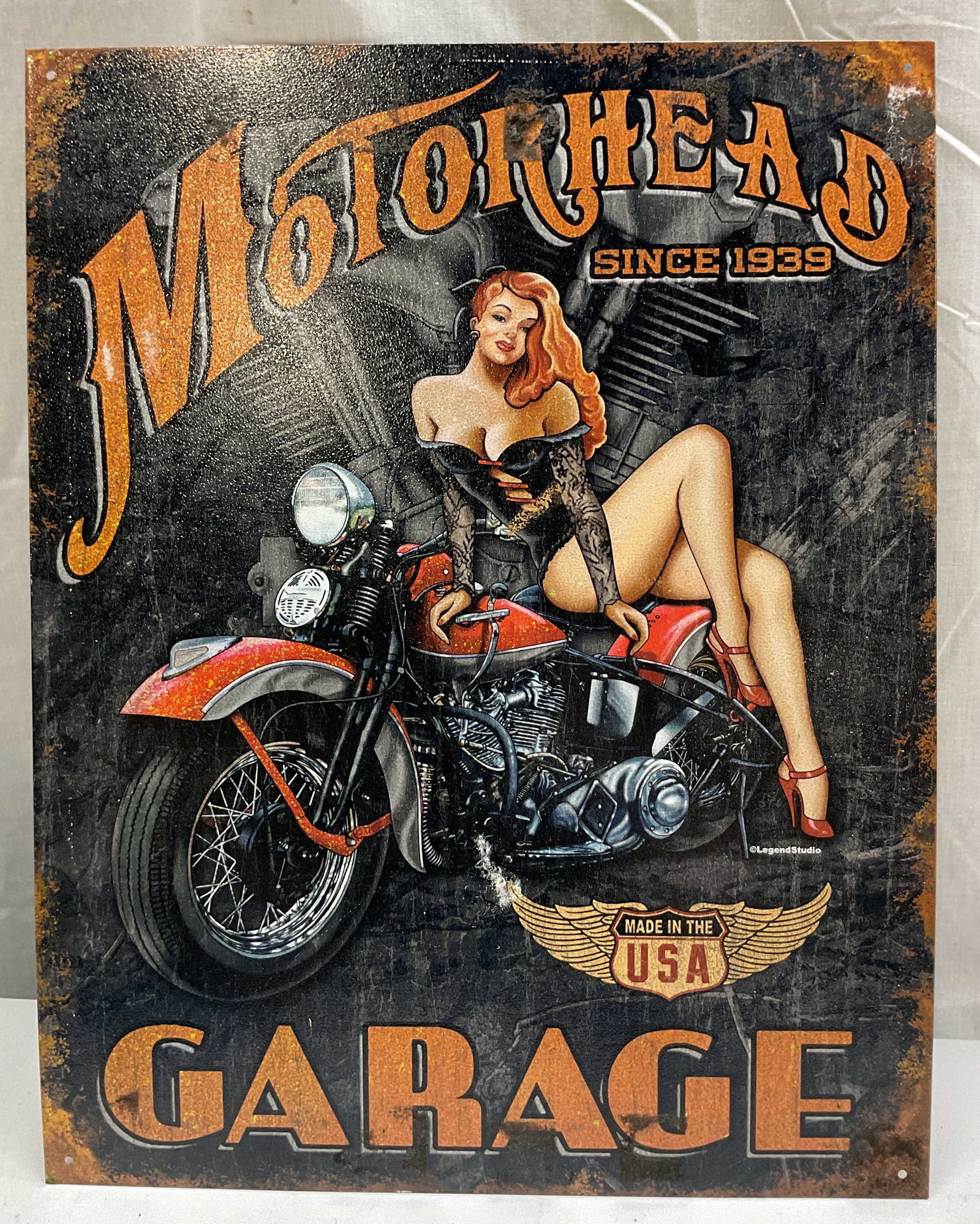 Legend Studio Motorhead Garage Wall Sign USA: Legend Studio Motorhead Garage tin wall sign. Made in USA. Copyright Legend Studio. Produced by Desperate Enterprises. Black and orange illustration of a motorcycle with a pin up model leaning against