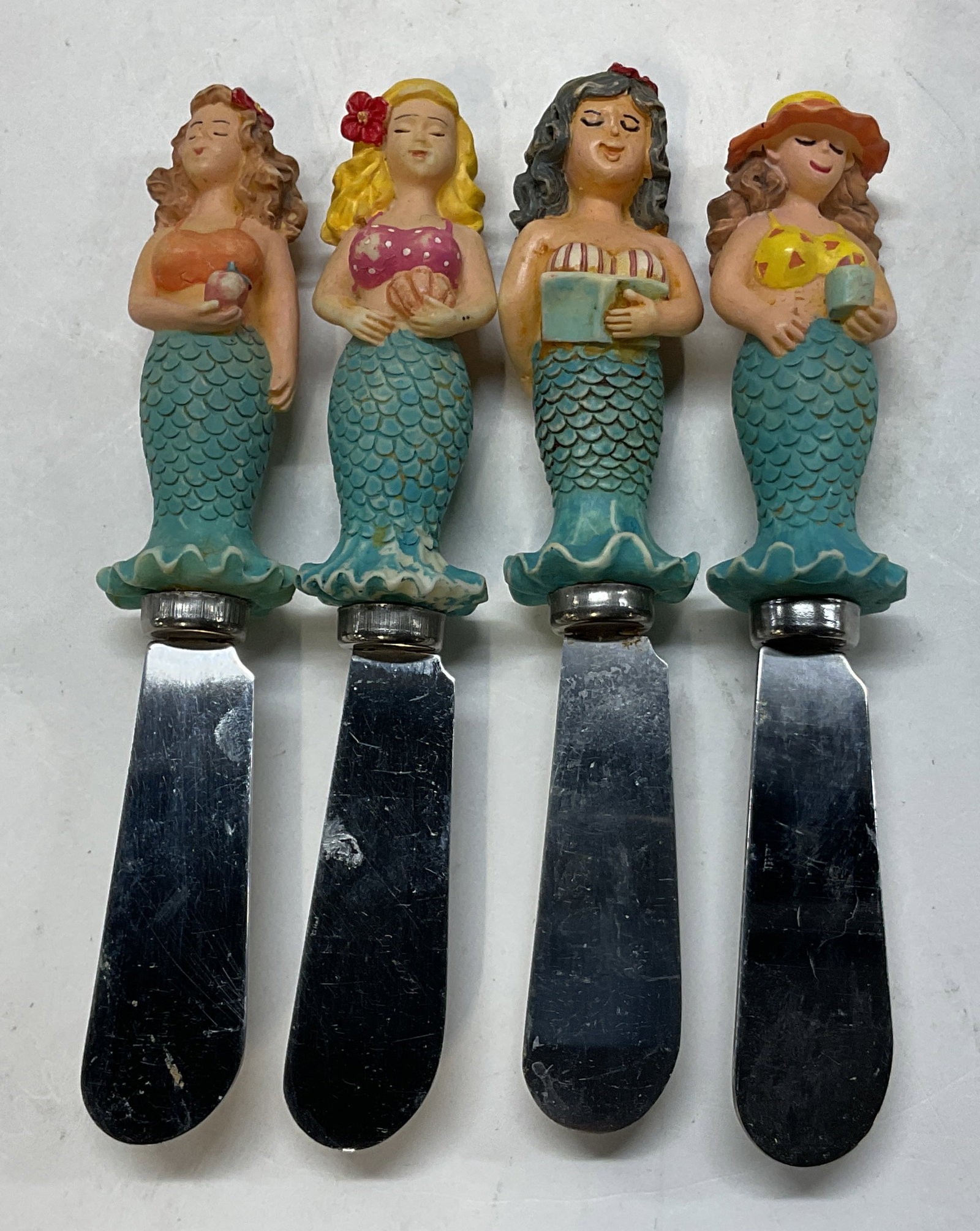 Lot 4 Stless Steel & Resin Mermaid Butter Knives: Lot of 4 stainless steel and resin mermaid butter knives. Marked Stainless Steel. Mermaid figures as the handles. They are holding objects such as a book, apple, etc. Hand painted in varying colors. D
