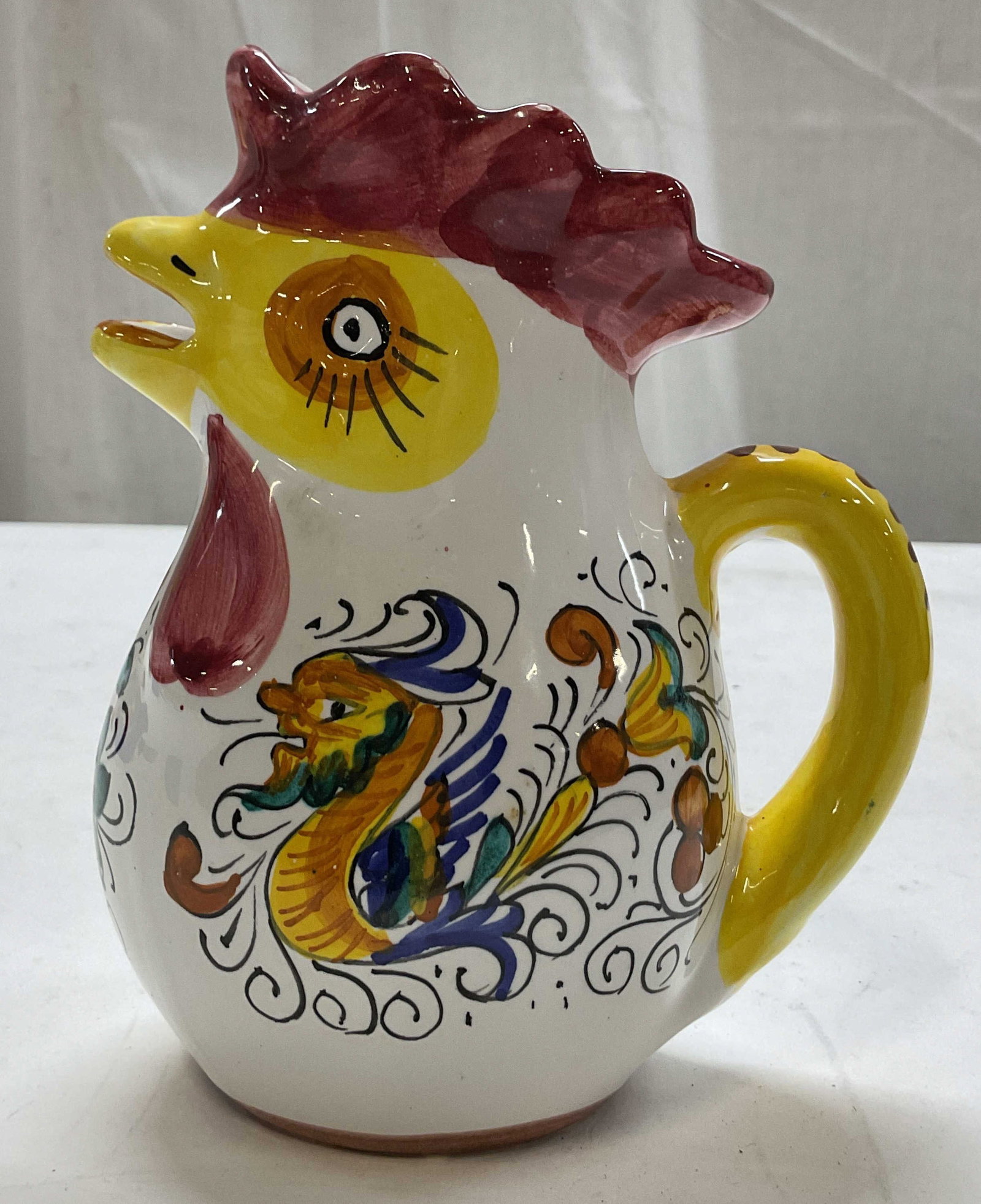 Williams Sonoma Ceramic Rooster Pitcher Italy: Williams Sonoma ceramic rooster pitcher. Marked on underside, made in Italy. Scalloped edge. Curved handle and spout is the mouth of the rooster. Hand painted features with floral and animal motifs.