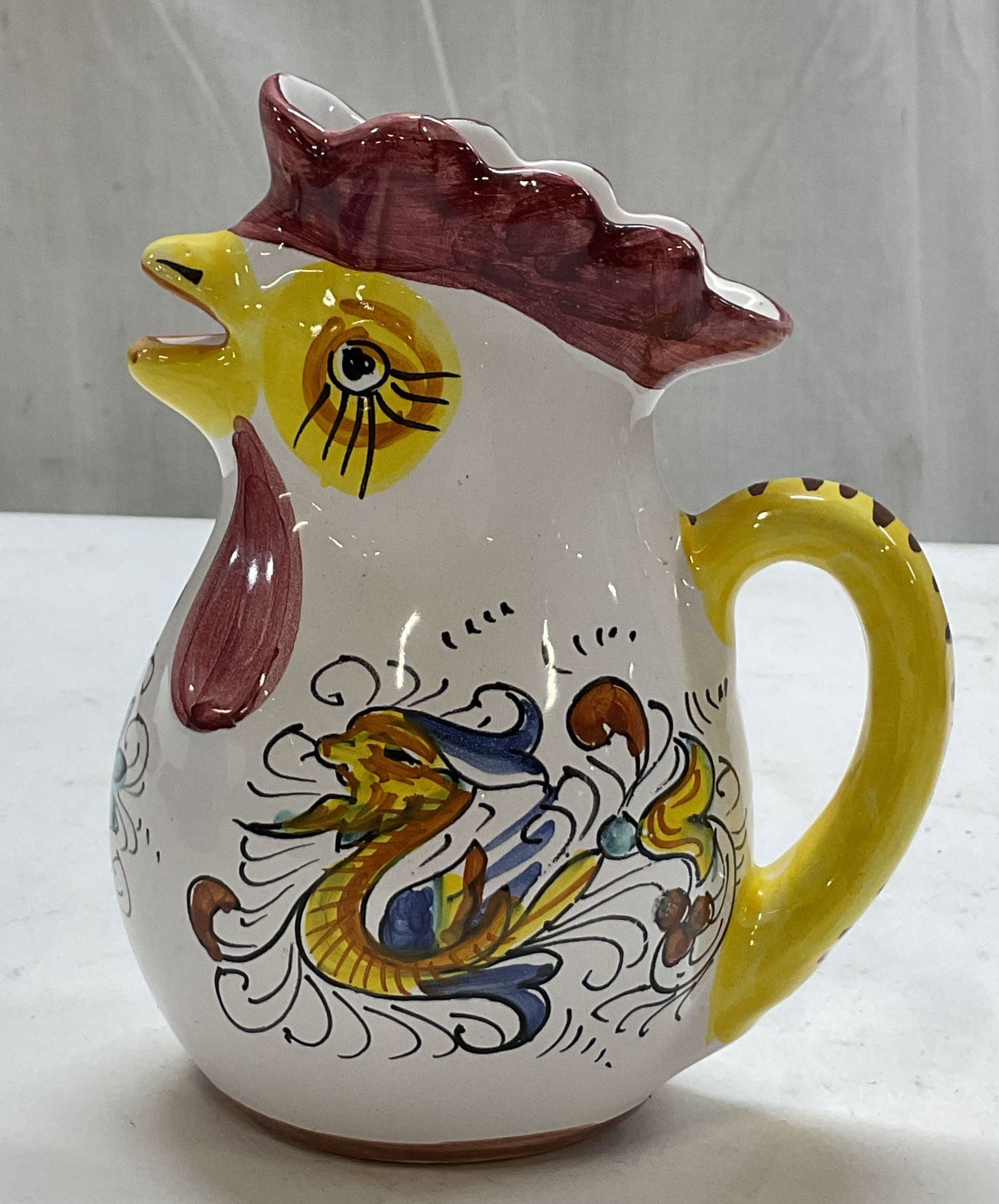 Williams Sonoma Ceramic Rooster Pitcher Italy: Williams Sonoma ceramic rooster pitcher. Marked on underside, made in Italy. Scalloped edge. Curved handle and spout is the mouth of the rooster. Hand painted features with floral and animal motifs.
