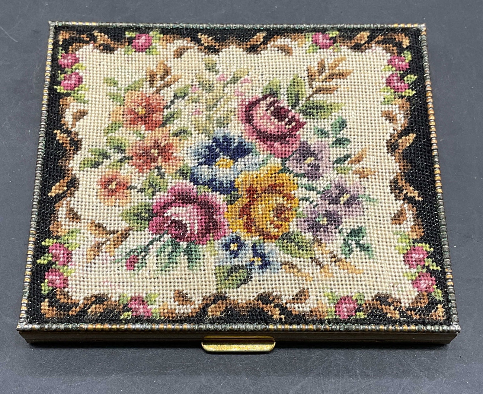 Vintage Needlepoint Flower Powder Compact: Vintage gold tone powder compact with needlepoint flower motif cover. Mirror and compartment on interior. Measures approx. 2.75 x 2.5 x 3/8 inches. Some scratching and discoloration. Needlepoint compa