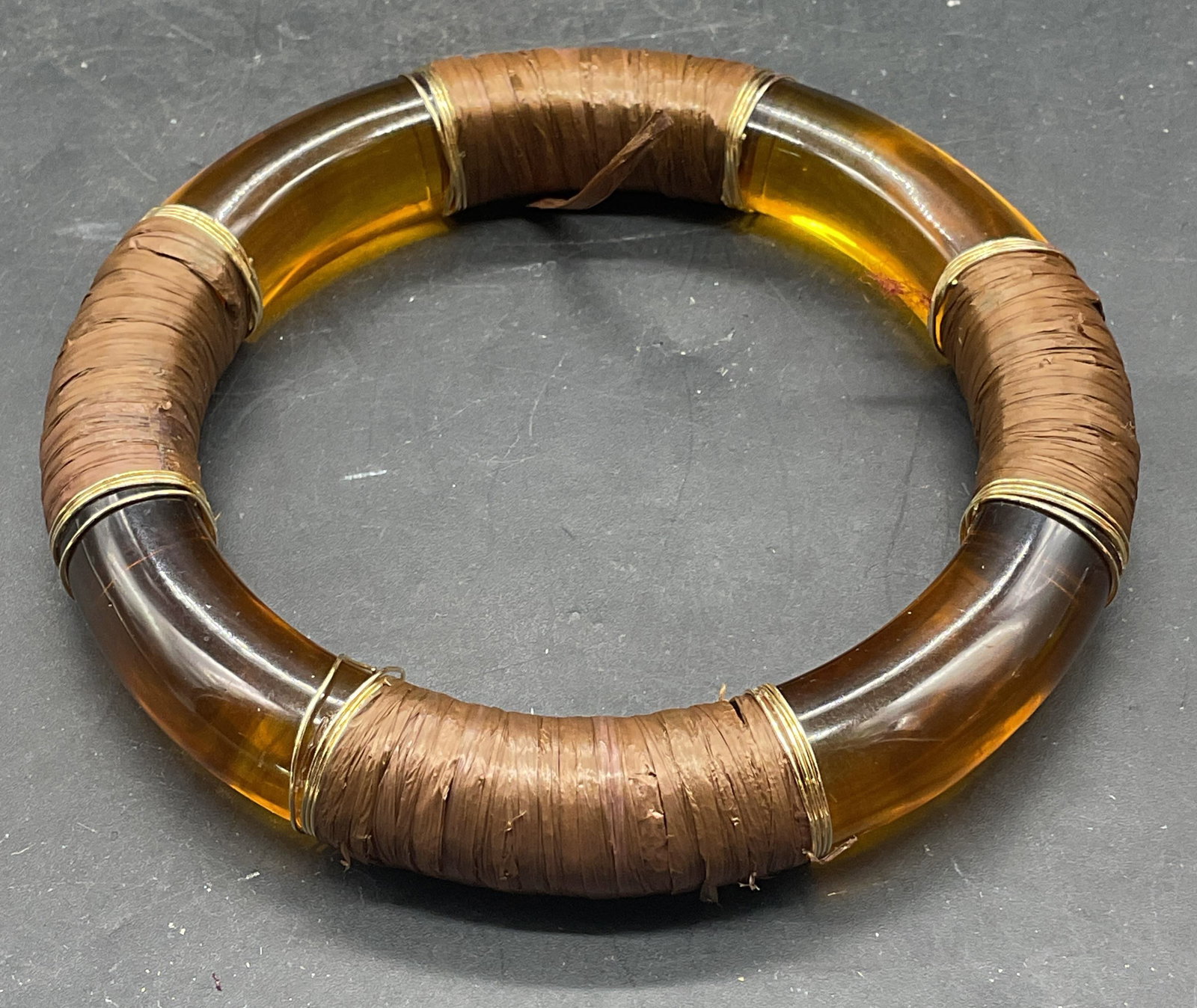 Amber Lucite Raffia Gold Tn Bangle Bracelet: Amber lucite bangle bracelet with brown raffia and gold tone wire wrapped decoration. Measures approx. 2.5 inches wide. Some scratching, warping to wire, tearing to raffia. Jewelry, estate jewelry, co