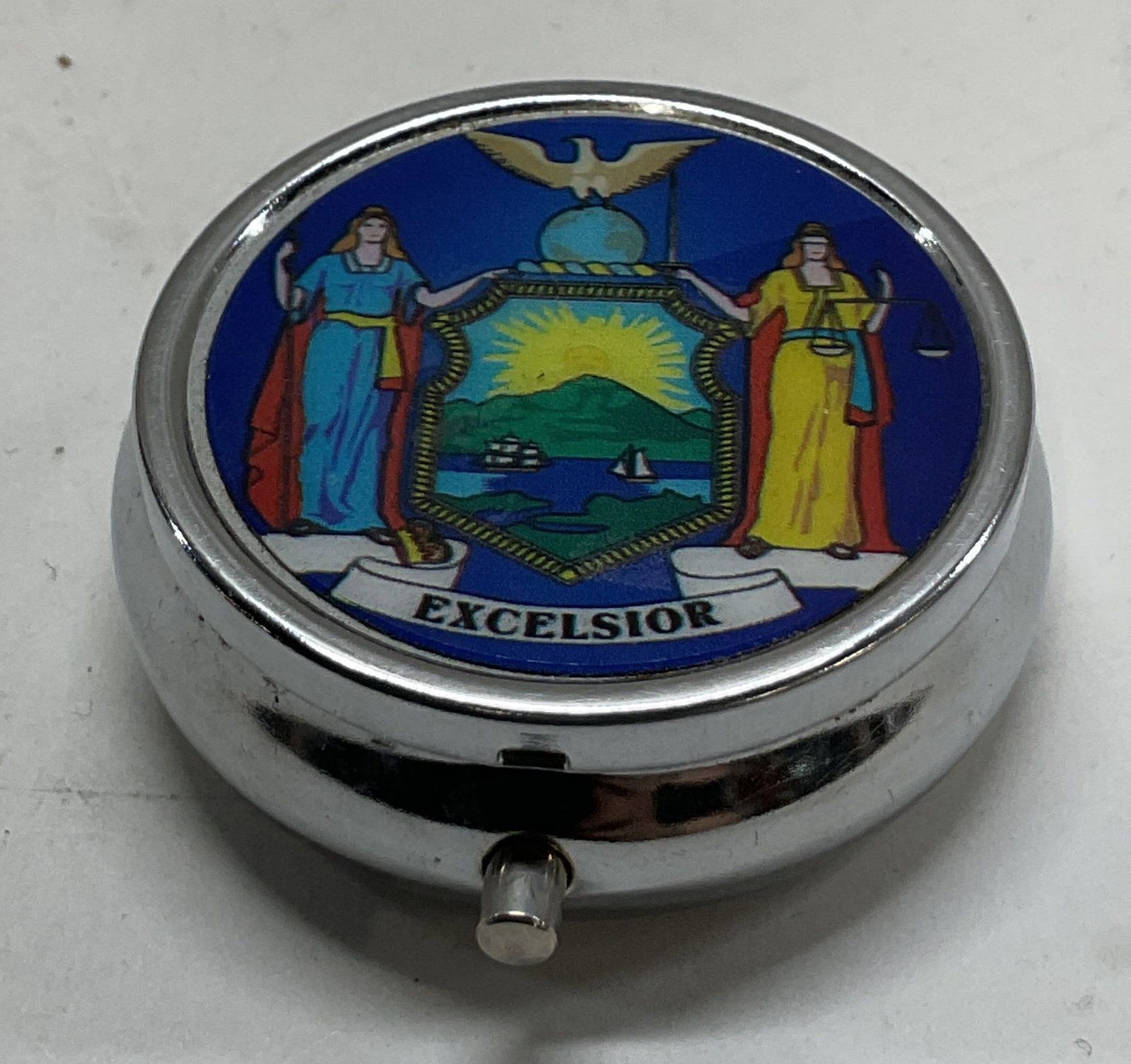 Vtg Silver Toned Metal NYS Coat Of Arms Pill Box: Vintage silver toned metal NYS Coat Of Arms hinged pill box. Round box with hinged lid. Press button opening box. Lid features the historical New York State coat of arms graphic with Liberty, Justice,