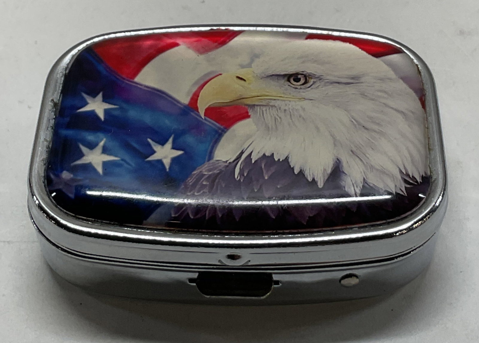 Vtg Silver Tned Metal Patriotic Bald Eagle Pill Bx: Vintage silver toned metal patriotic bald eagle hinged pill box. Oval rectangular shaped box with hinged lid. Press button opening box. Lid features a bald eagle with the American Flag in the backgrou