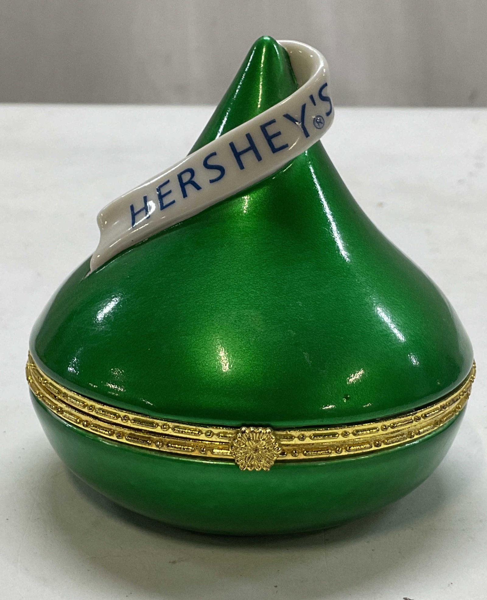 Vintage Green Porcelain Hersheys Kiss Trinket Box: Vintage green porcelain Hersheys kiss hinged trinket box. Box shaped like a Hersheys kiss. Bright green color. Hinged lid. Gold toned hardware with decorative floral motif closure and embossed scroll 