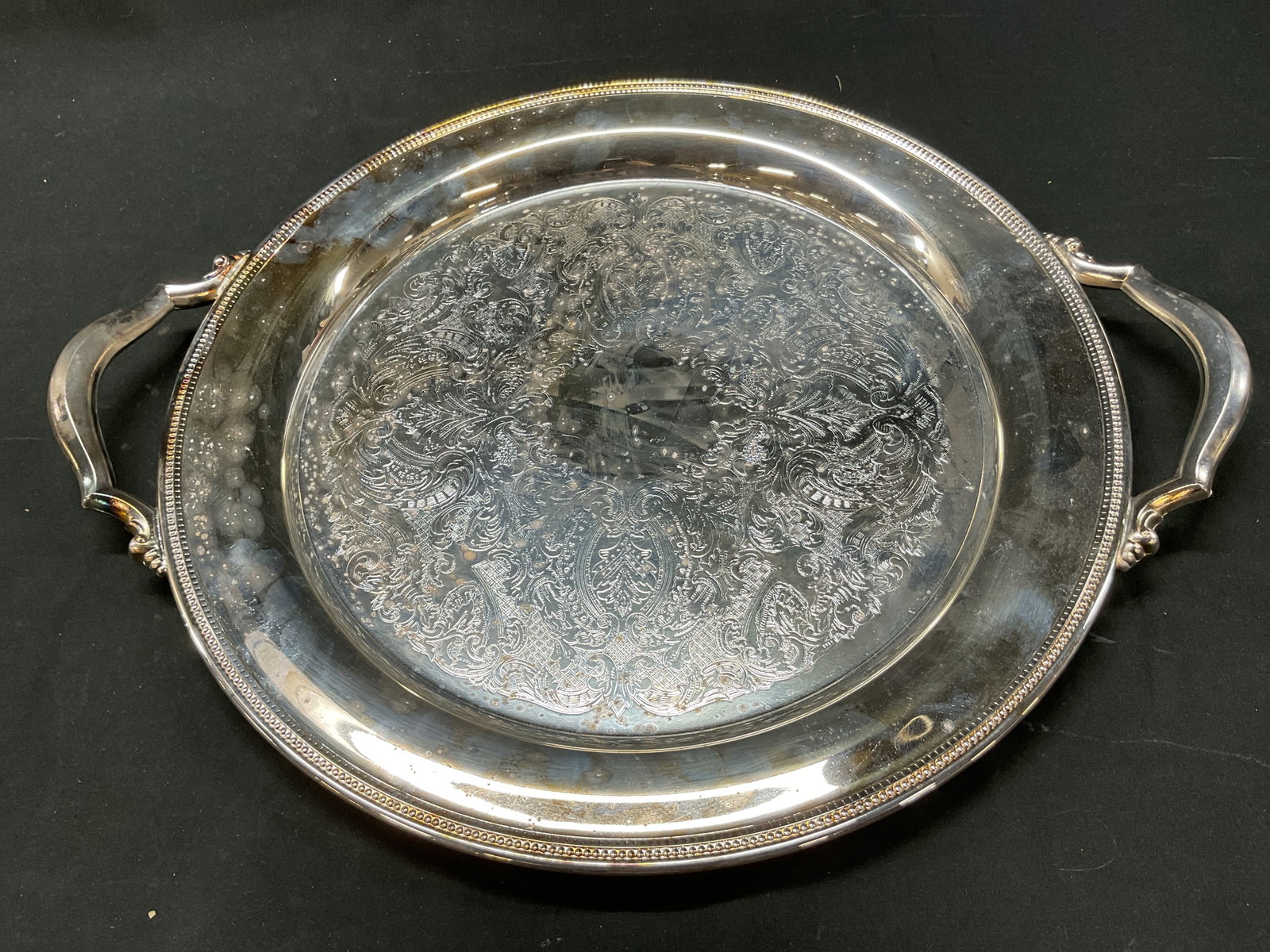 Oneida Silver Plated Handled Serving Tray USA: Oneida silver plated handled serving tray. Marked Oneida USA on underside. Ornate handles. Engraved floral and scroll motifs. Beaded rim. Measures approx 18 inches in diameter. Scratching and wear to