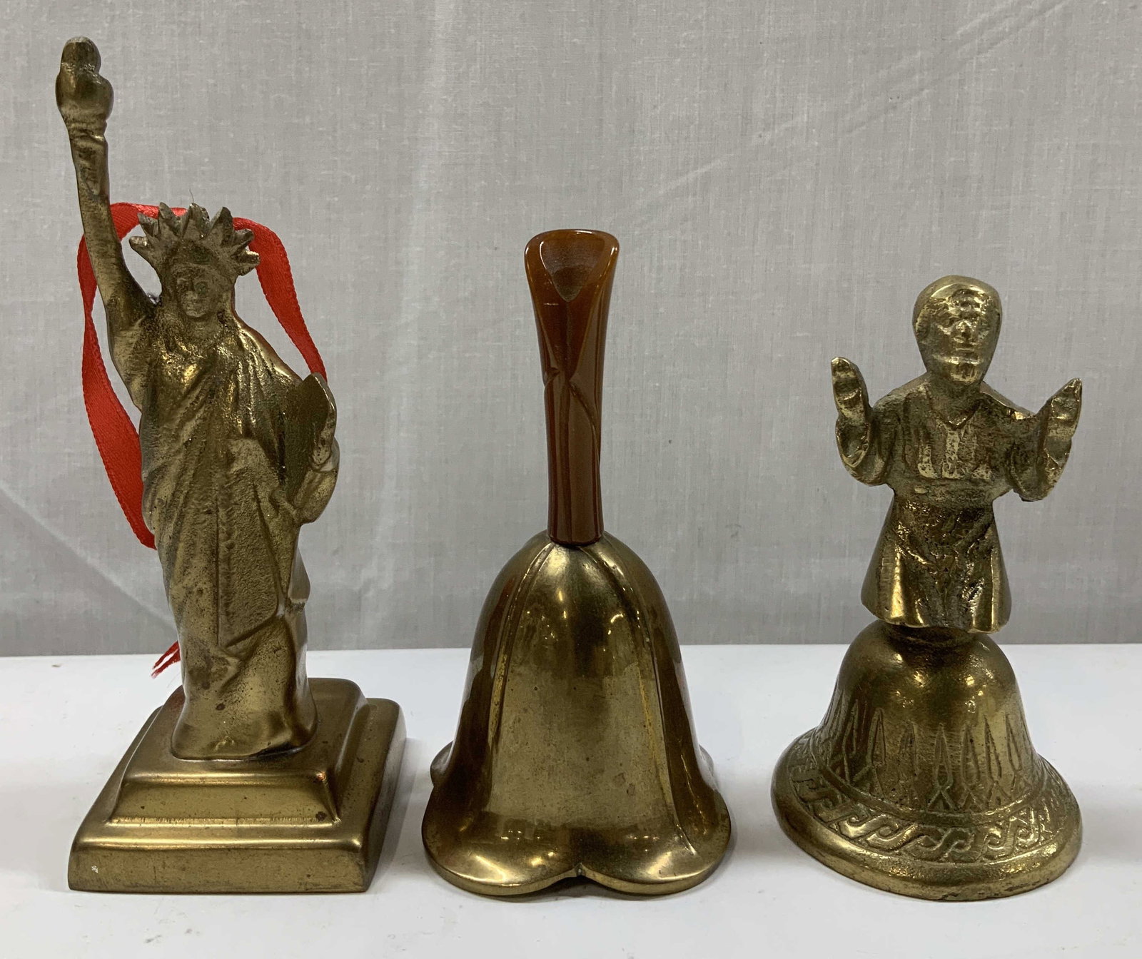 Lot 3 Vtg Brass Bells & Ornament RESTORATION INDIA: Lot includes, 1 brass bell with a bakelite handle, 1 brass bell featuring a figural handle of a kneeling man in prayer & 1 Statue of Liberty ornament. Signed & marked on underside. Reads, RESTORATION