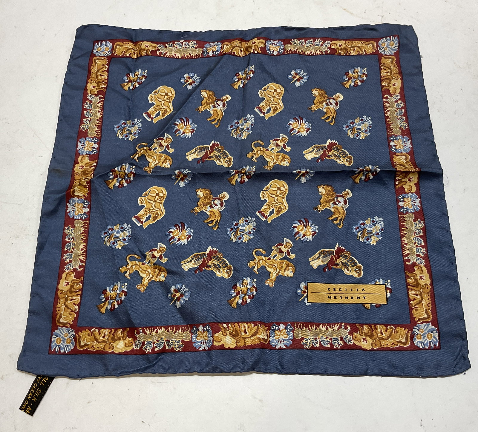 Cecilia Metheny Blue Silk Pocket Square Italy: Cecilia Metheny blue silk pocket square. Made in Italy. Blue background. Animal, figural, and floral motifs in cream, red, blue. Decorative red border. Measures approx 12 x 12 inches. Blemishes. Prope