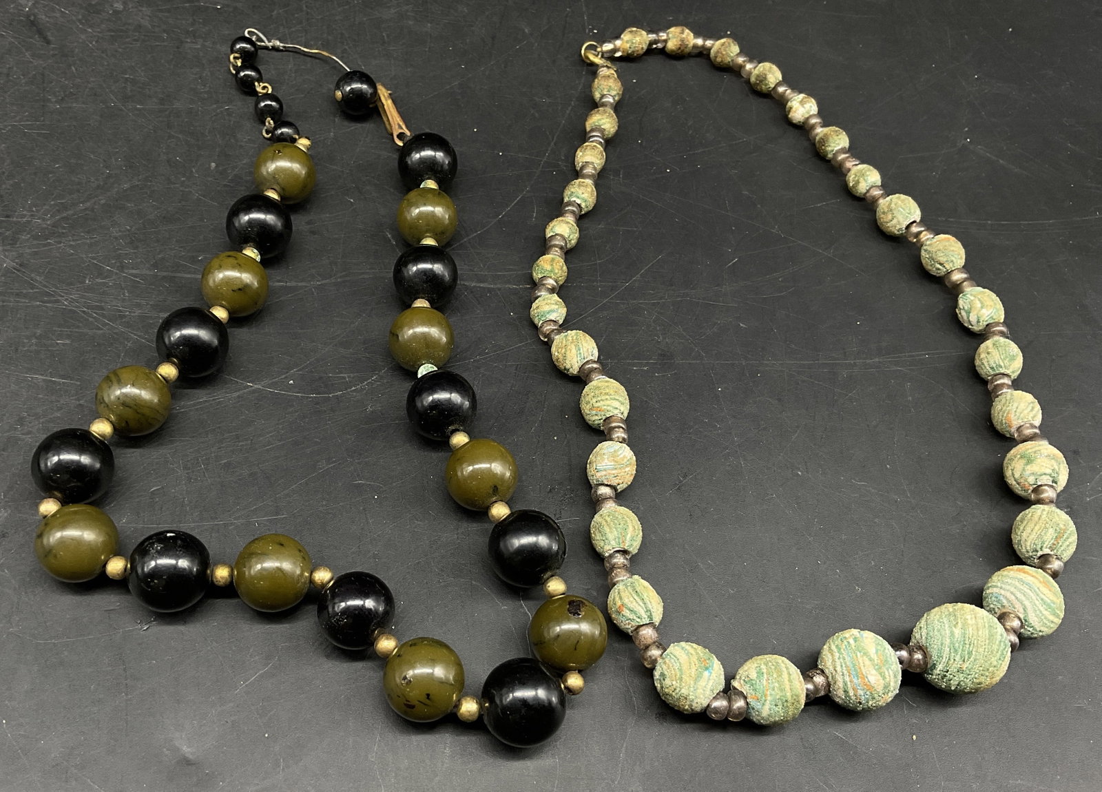 Lt2 Earthy Green Beaded Necklaces: 2 Earthy Green Beaded Necklaces. Includes Black and Olive Green Resin Beads with Gold-Toned Spacer Beads Necklace and a Green Swirled Wooden Beaded Necklace. Corrosion and Aging to Metal Present. Meas
