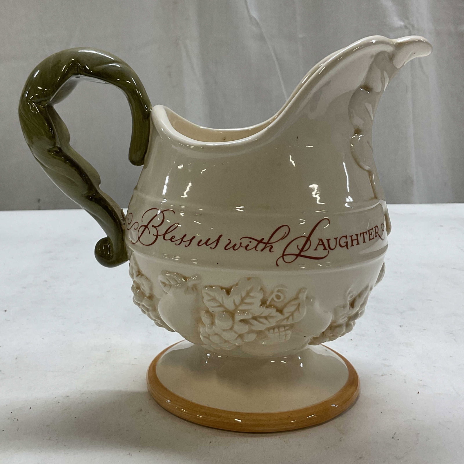 Vintage Decorative Ceramic Fall Themed Pitcher: Vintage decorative ceramic fall themed pitcher. Round pedestal base with orange rim. Green leaf motif handle. Embossed acorn, fruit, leaf motifs. Curved spout. Surface reads Bless Us With Laughter