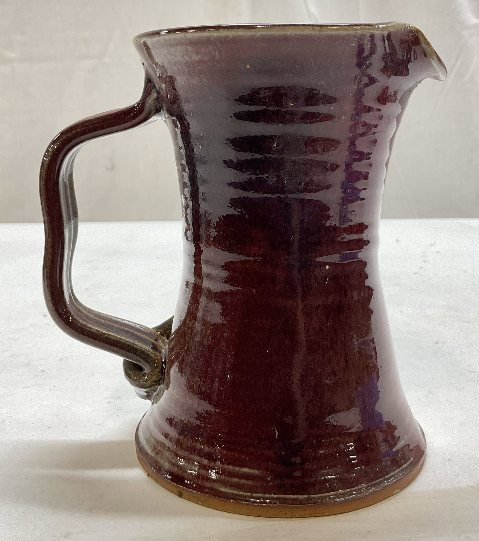 Vintage Red Glazed Pottery Pitcher: Vintage red glazed pottery pitcher. Signed on underside, reads illegible. Curved spout. Scalloped, ribbed, and curved handle. Flared round base. Ribbed body, cylindrical shape. Glossy finish, red