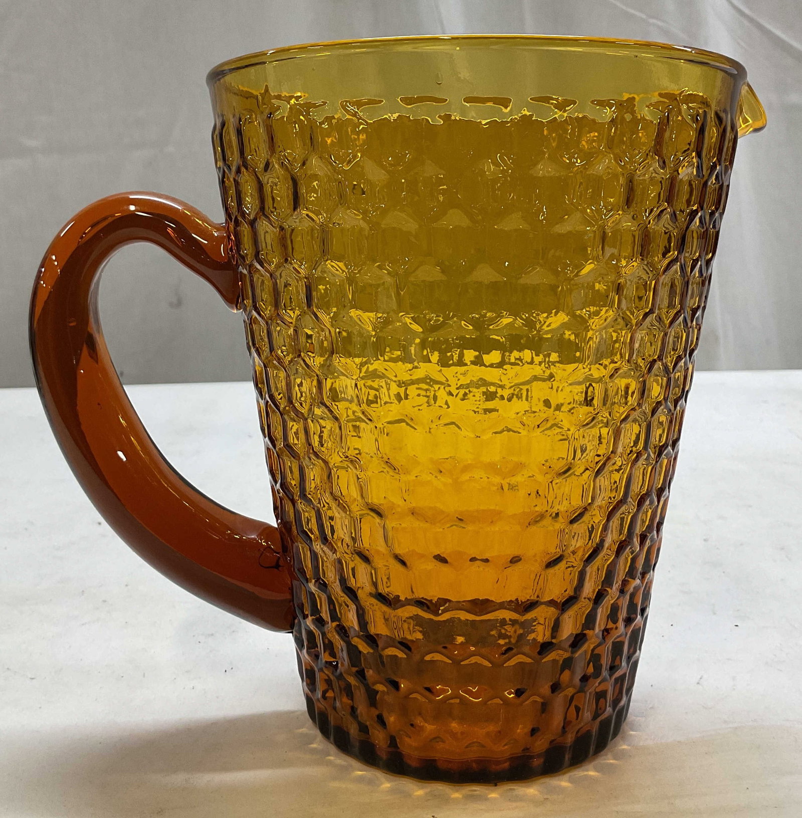 Vintage Amber Glass Pitcher: Vintage amber glass pitcher. Tall cylinder shape, bulbous body. Curved handle and spout. Textured geometric pattern. Measures ht approx 7 inches diam approx 5.5 inches. Scratching. Pitcher, home