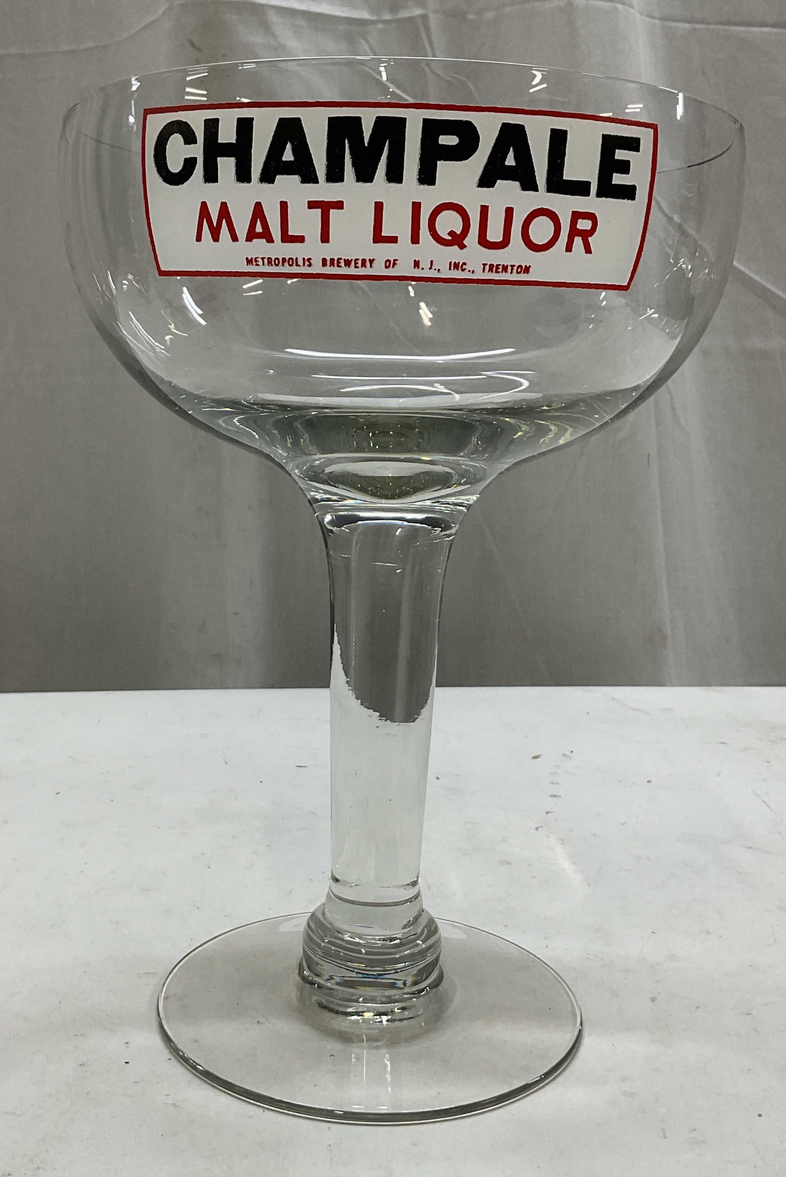 Vtg Oversized Champale Malt Liquor Display Goblet: Vintage oversize Champale Malt Liquor glass display goblet. Round base and thick stem. Wide bowl and rim. Features the iconic red and white Metropolis Brewery logo. Surface reads Champale Malt Liquor