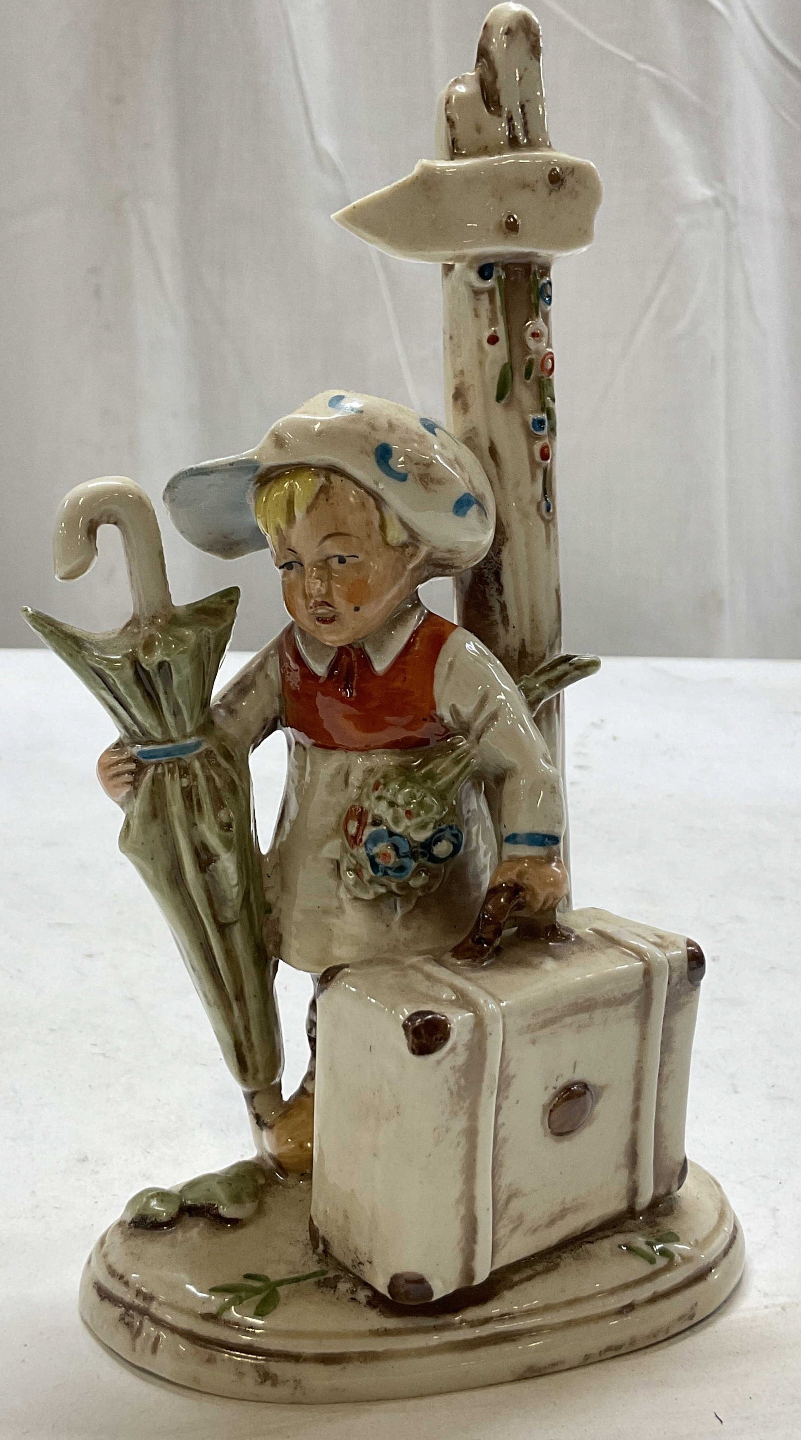Vintage Porcelain Traveler Figure Germany: Vintage porcelain traveler figure. Marked Germany on underside. Oval base. Young figure wearing a hat carrying flowers, an umbrella, and a suitcase standing next to a signpost. Varying colors and