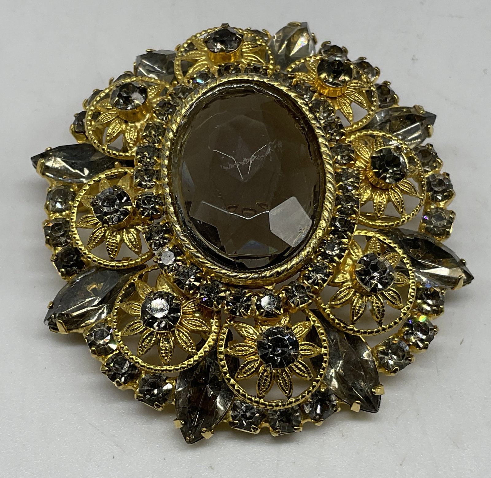 VTG Gold-Toned Filigree Statement Brooch: Vintage Gold-Toned Filigree Statement Brooch. Floral designs. smoky domed oval glass center. smoky teardrop and circular rhinestone accents. Condition consistent with exposure. Measures app 2in Diam.