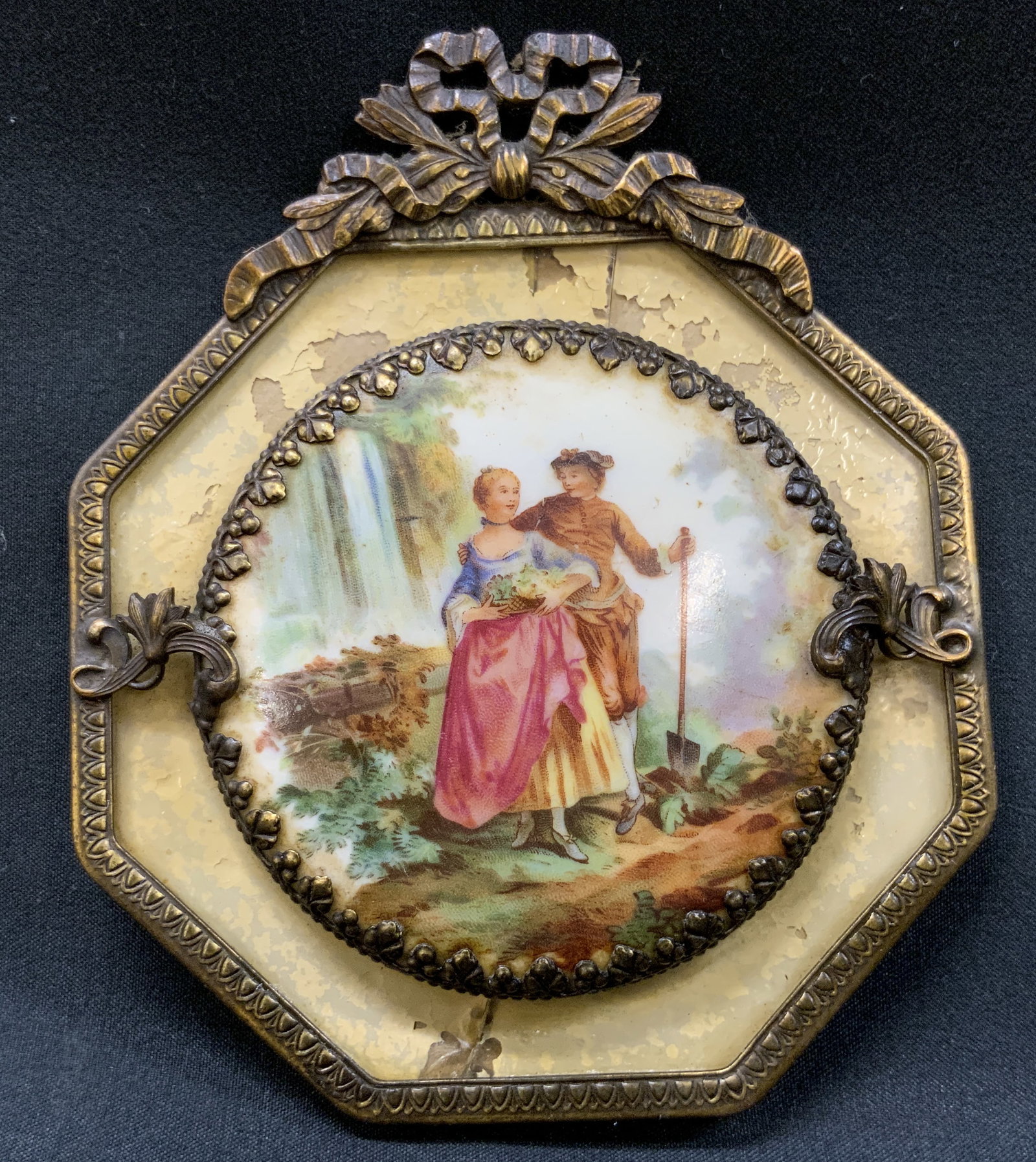 Vtg Courting Couple Framed Mini Wall Art: Vintage octagonal portrait plaque features central circular celluloid disc with courting couple in landscape setting imagery. Secured in decorative metal border ring with side leaf tabs mounted onto c
