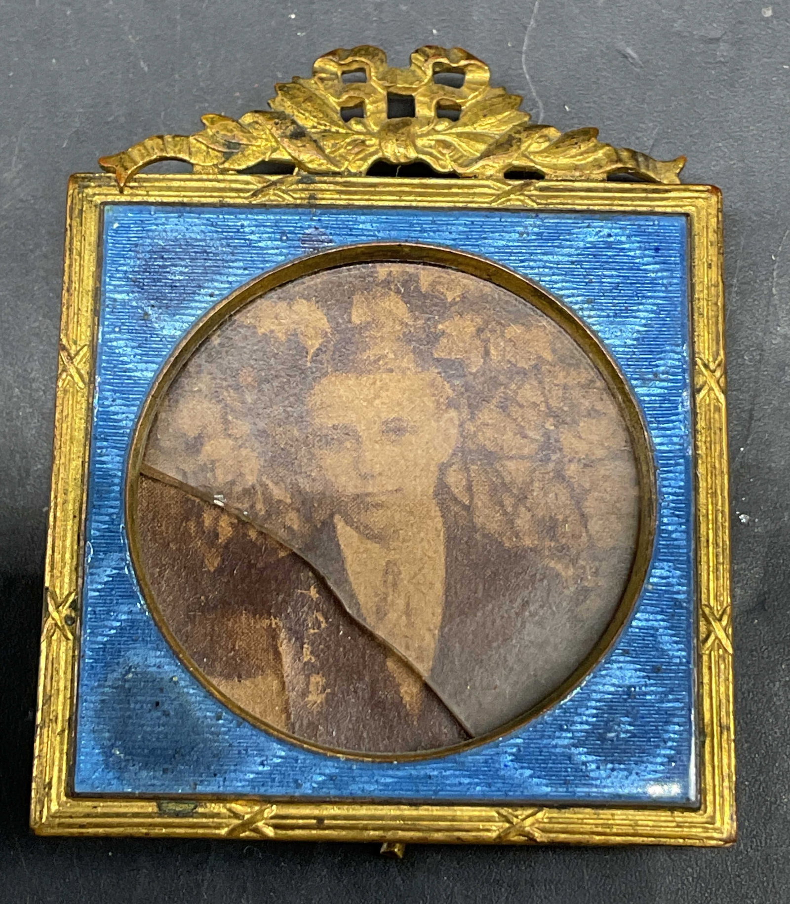 Antique Brass Blue Enamel Frame Czechoslovakia: Antique brass blue enamel picture frame. Backside is marked Made In CzechSlov. Square frame with ornate bow accent. Blue enamel inner frame. Measures approx 1.5 x 2 inches. Interior glass is cracked,