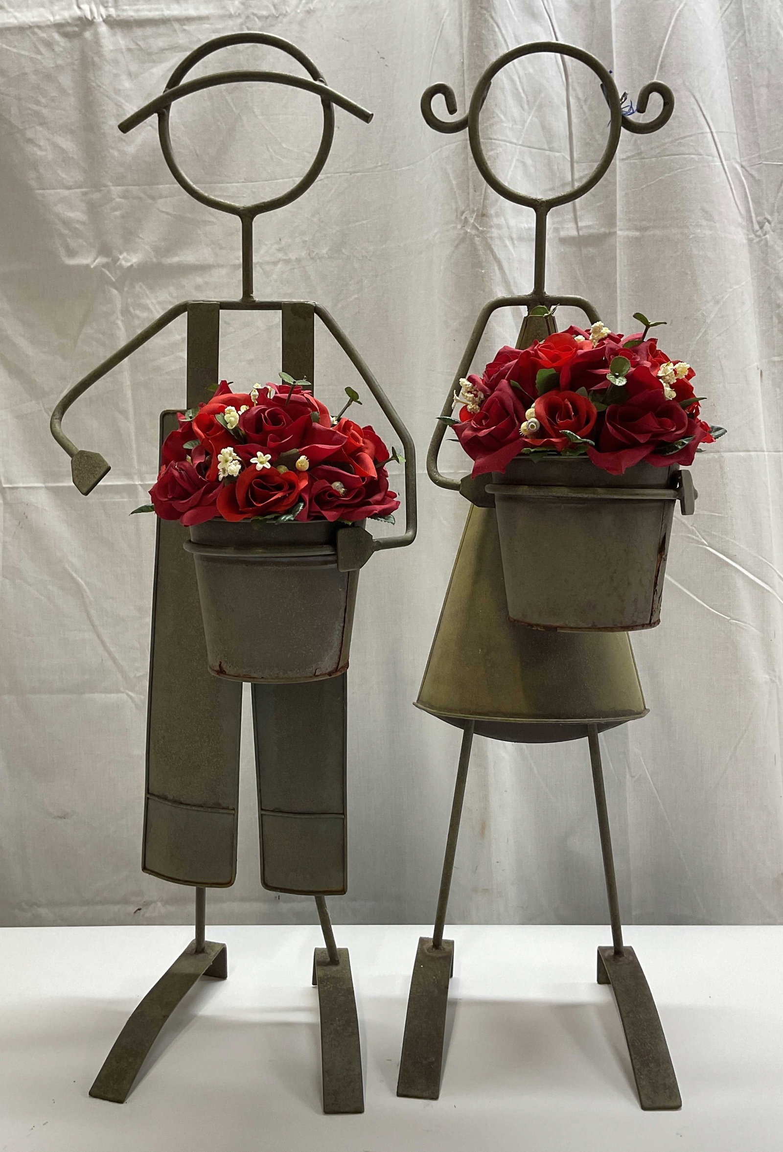 Pair Vtg Sheet Metal Stick Figure Garden Statues: Pair of vintage sheet metal stick figure garden statues. Boy and girl stylized stick figure design. They are both holding buckets with red faux flowers. Sheet metal, possibly iron or steel. Measures h