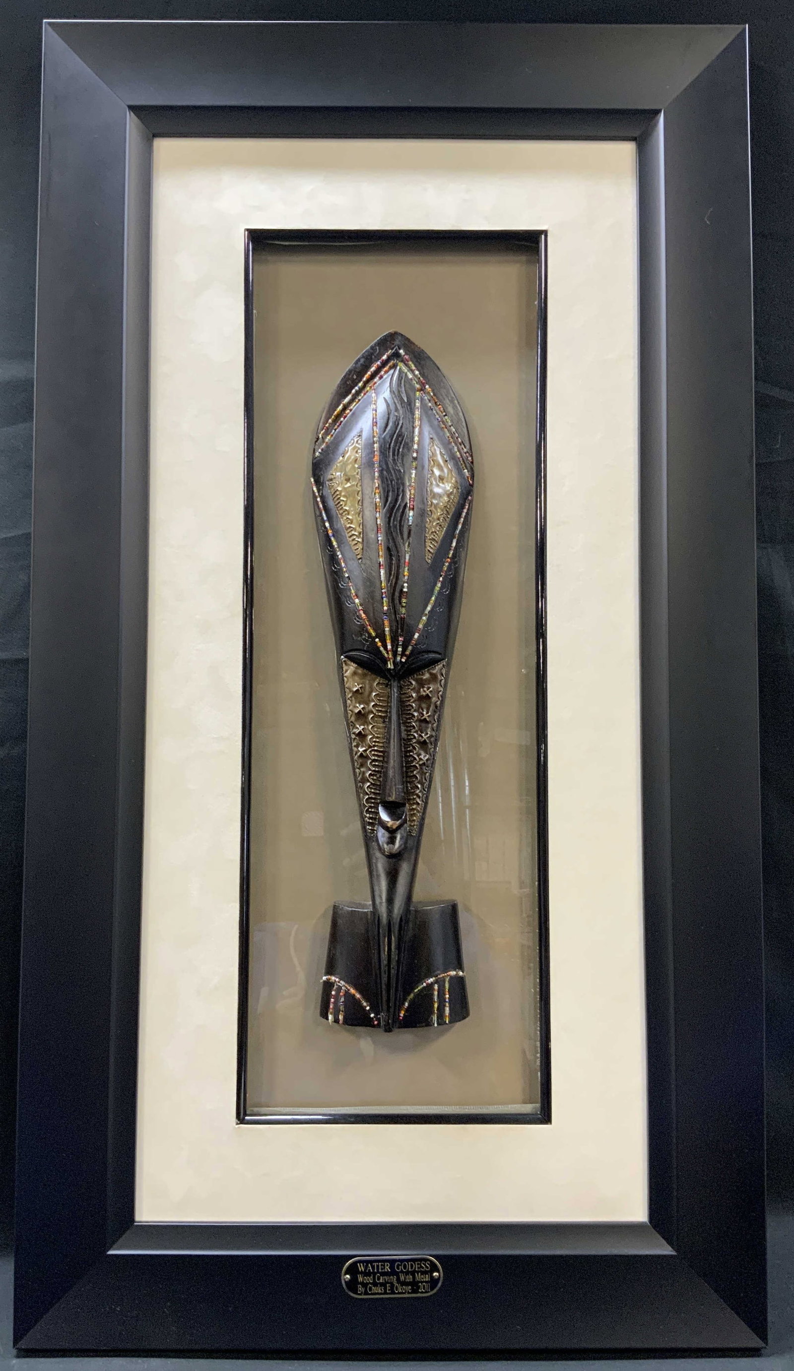 Chuks E. Okoye Wood Sculpture, Framed: Chuks E. Okoye framed three dimensional wood carving with metal wall sculpture. Titled Water Goddess, made in 2011. Plaque with title, medium, and artist name on bottom of frame. Frame, surface wear t