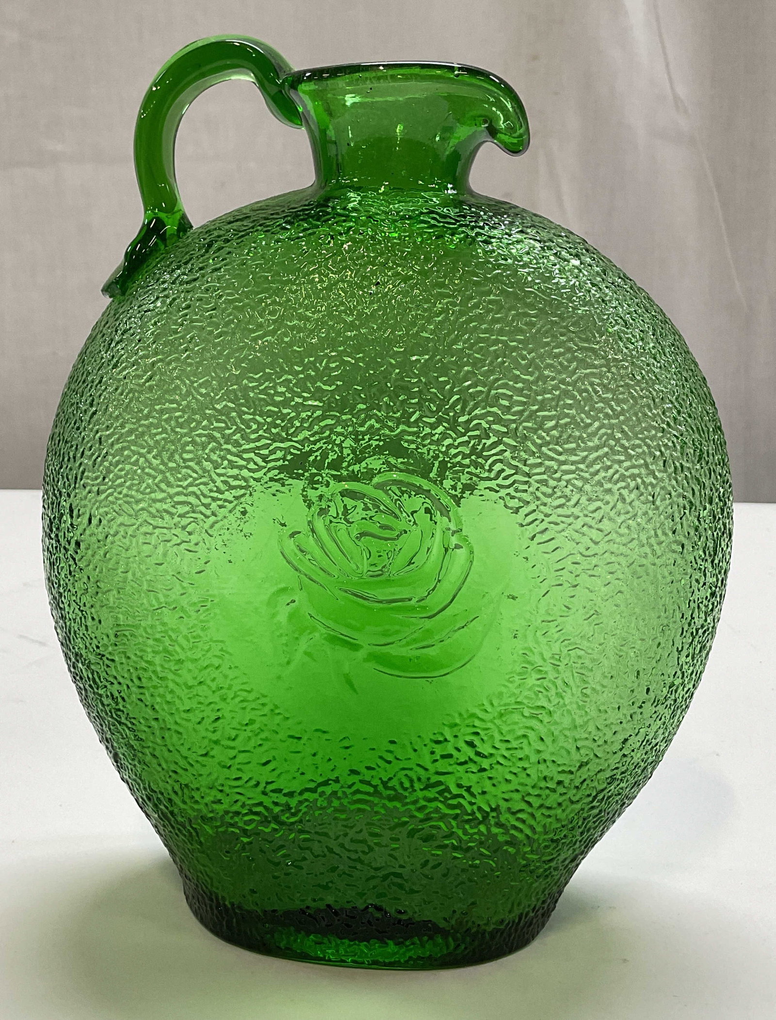 Antq Hand Blown Green Art Glass Jug: Antique hand blown green art glass jug. Pontil on underside, hand crafted. Bulbous body. Applied handle and curved spout. Textured body and rose motif in center. Measures ht approx 7.5 inches diam