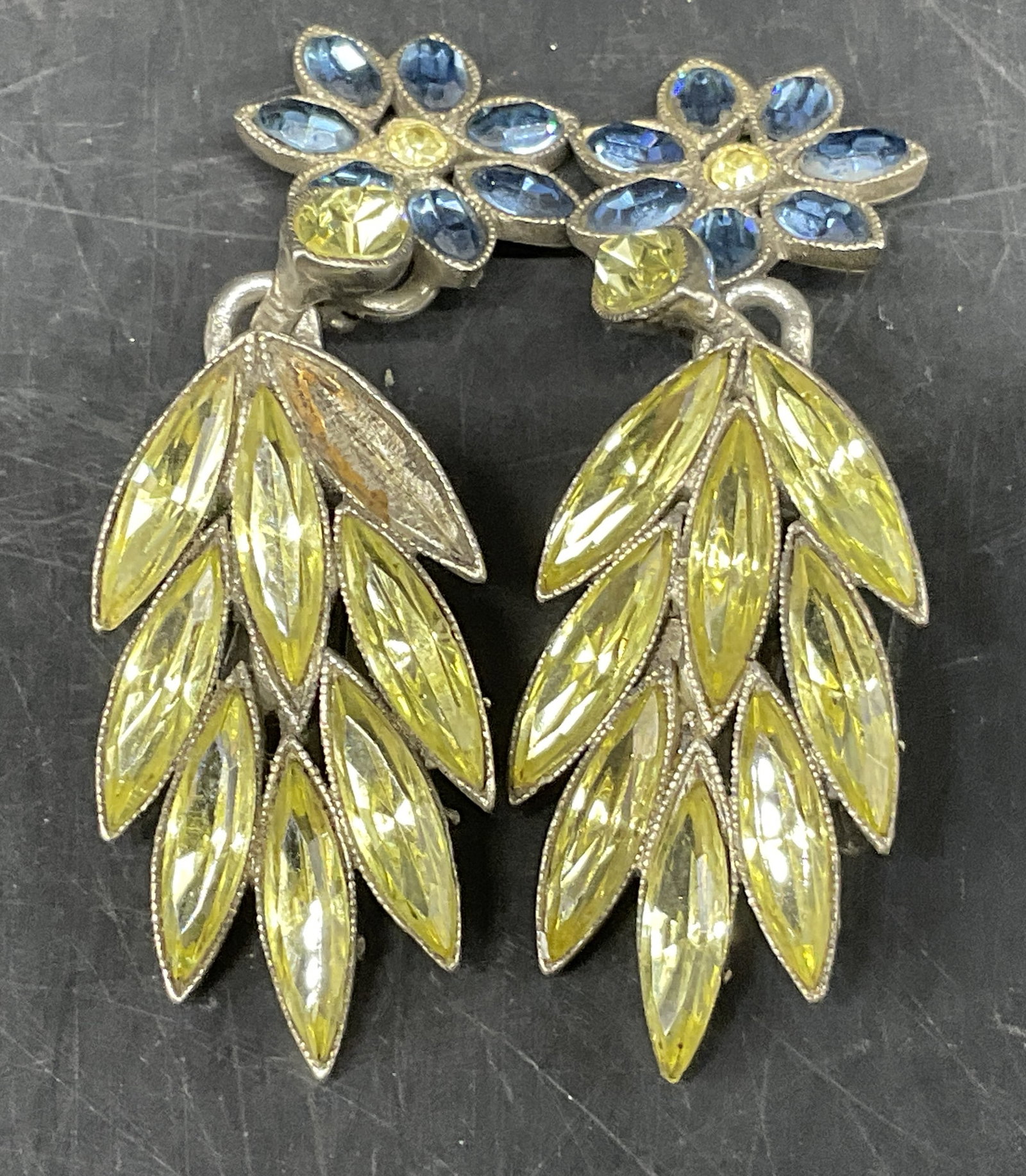 VTG Roxanne Assoulin Floral Clip-On Earrings: Vintage Roxanne Assoulin Silver-Toned Clip-On Earrings. Blue flower design with yellow center and dangling navette rhinestone cluster. Missing stone. Measures approx 2.5in H x 0.75in W. vintage