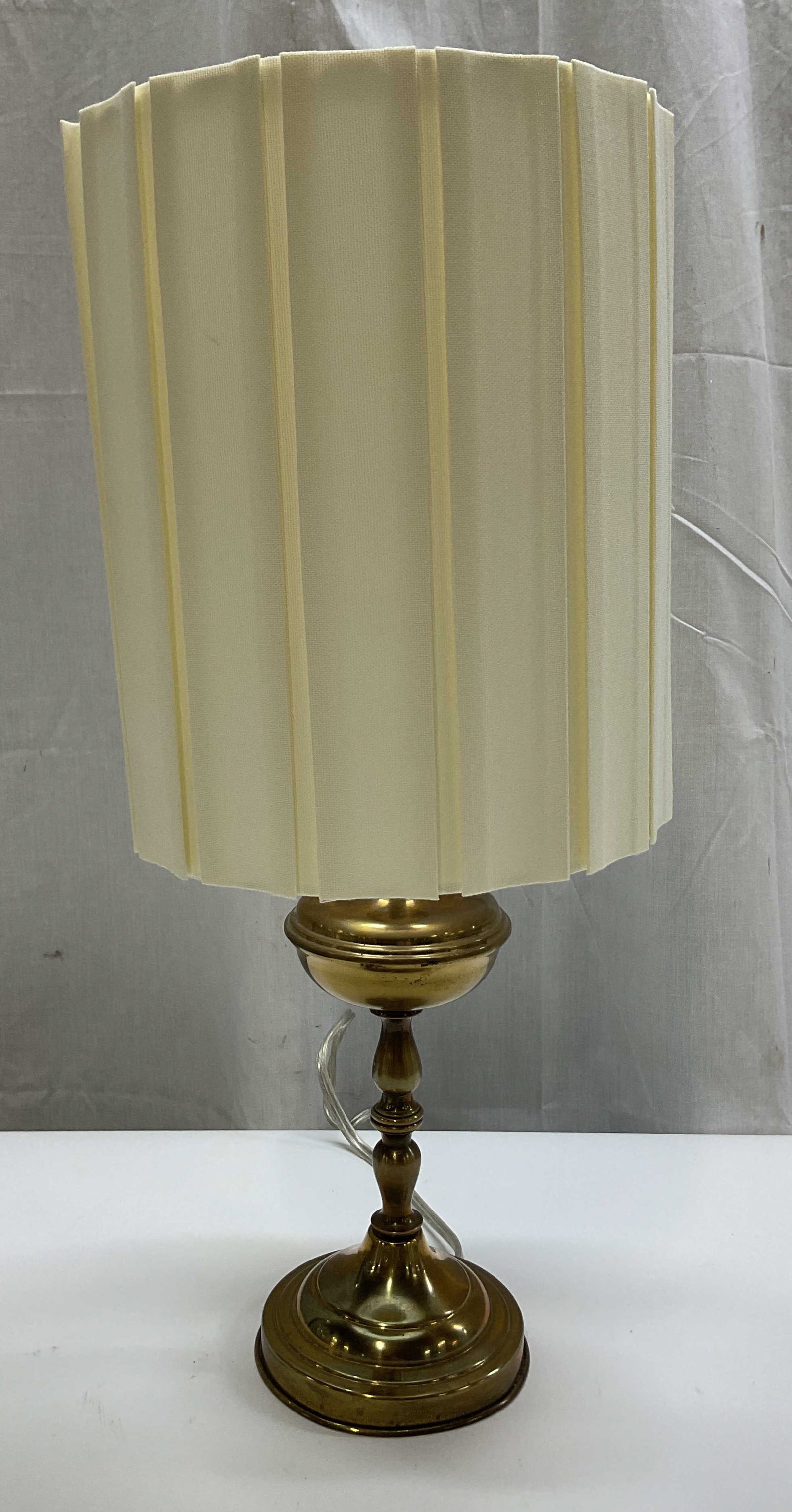Vintage Brass Table Lamp W Shade: Vintage brass table lamp. Comes with pleated fabric cream drum shade. Round pedestal base. Multi tiered turned column. Decorative finial. Measures ht approx 20 inches diam approx 5.5 inches. Not teste
