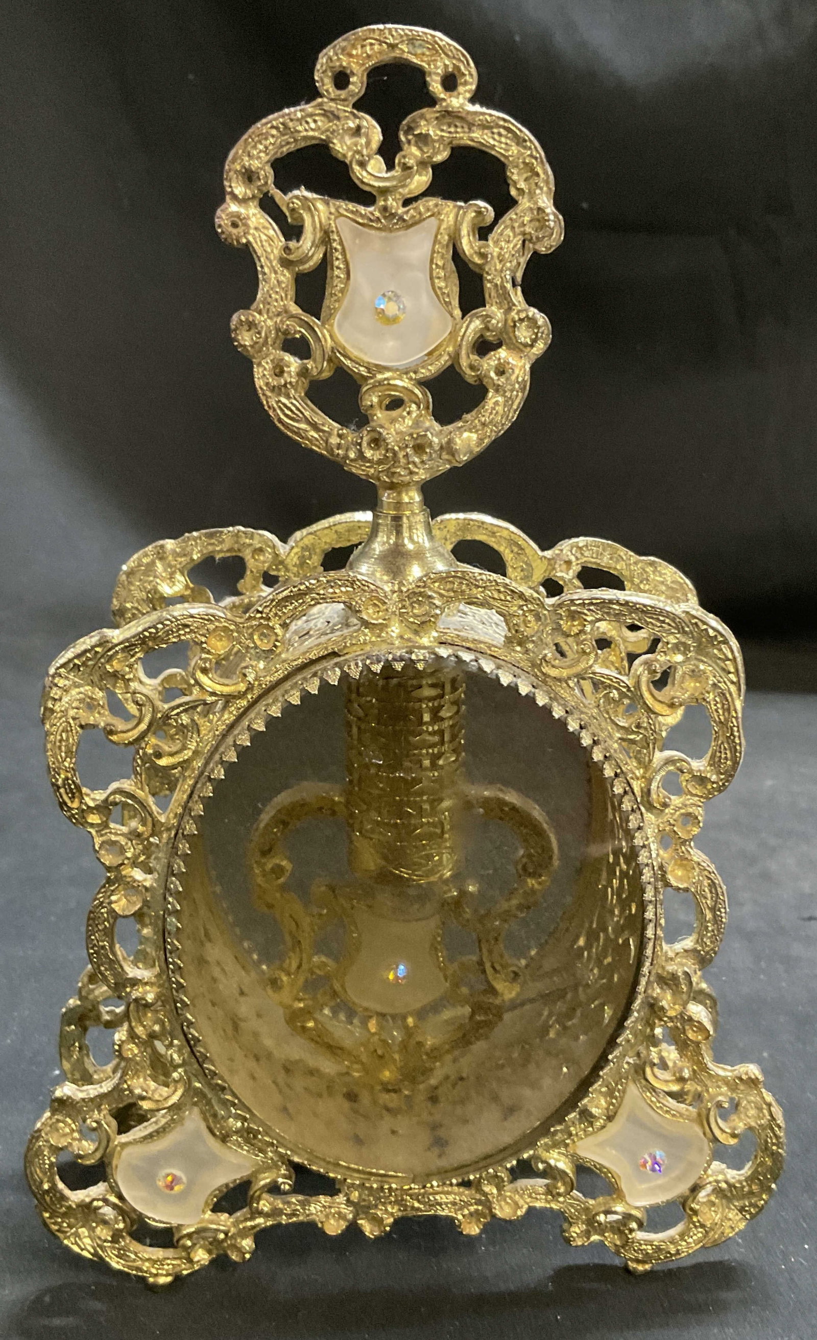 VTG Hollywood Regency Gilt Filigree Perfume Jacket: Vintage Hollywood Regency Gilt Filigree Perfume Jacket. Smoky grey glass oval panel. White glass accents with rhinestones. Detached glass piece inside. Missing rhinestone. Measures approx 9.5in H x 4.
