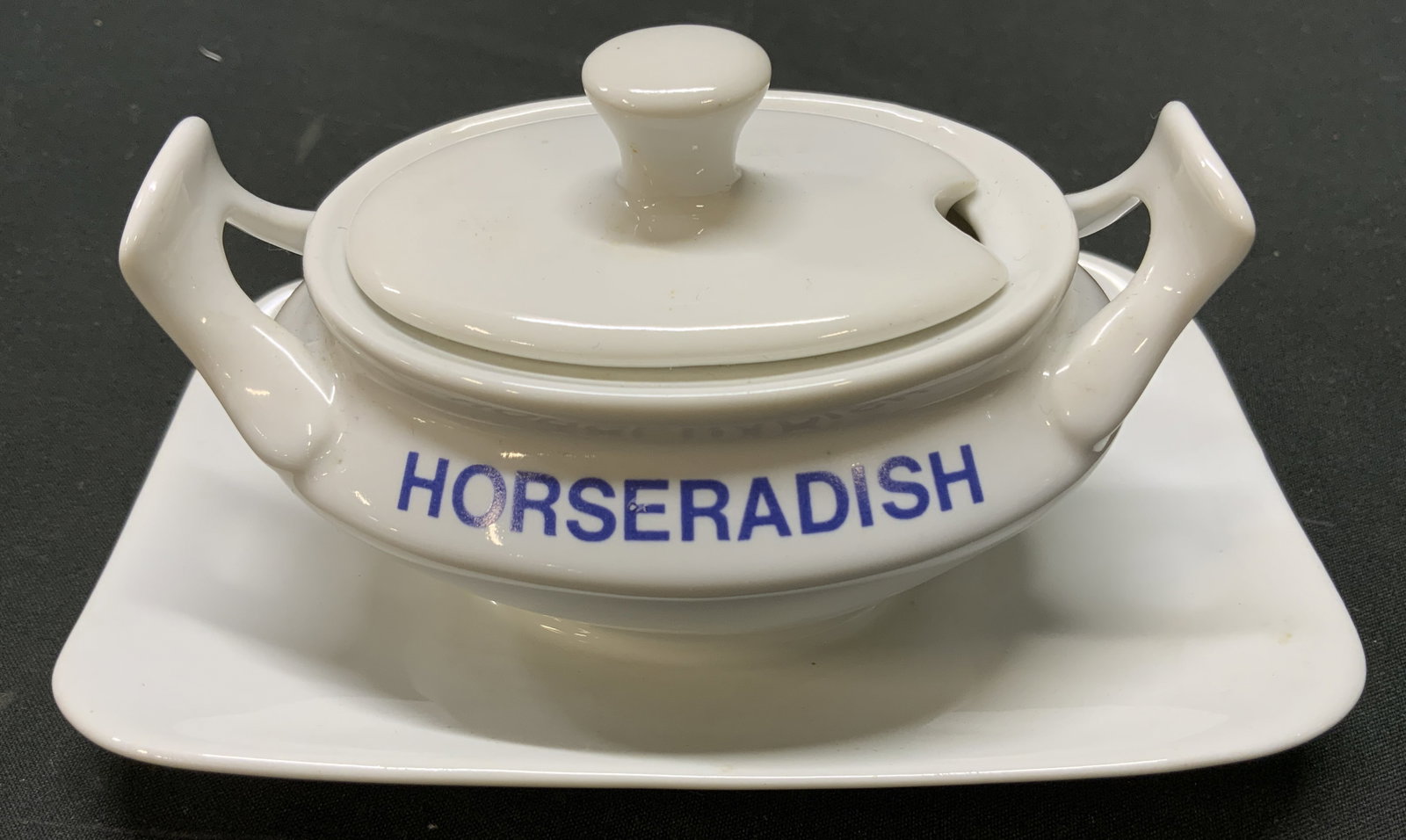 Vtg Porcelain Horseradish Dish Attached Underplate: Vintage white porcelain horseradish dish with built in handles, notched lid for serving spoon, integrated non removable underplate saucer. Stamped HORSERADISH in blue block lettering. Measures approx.