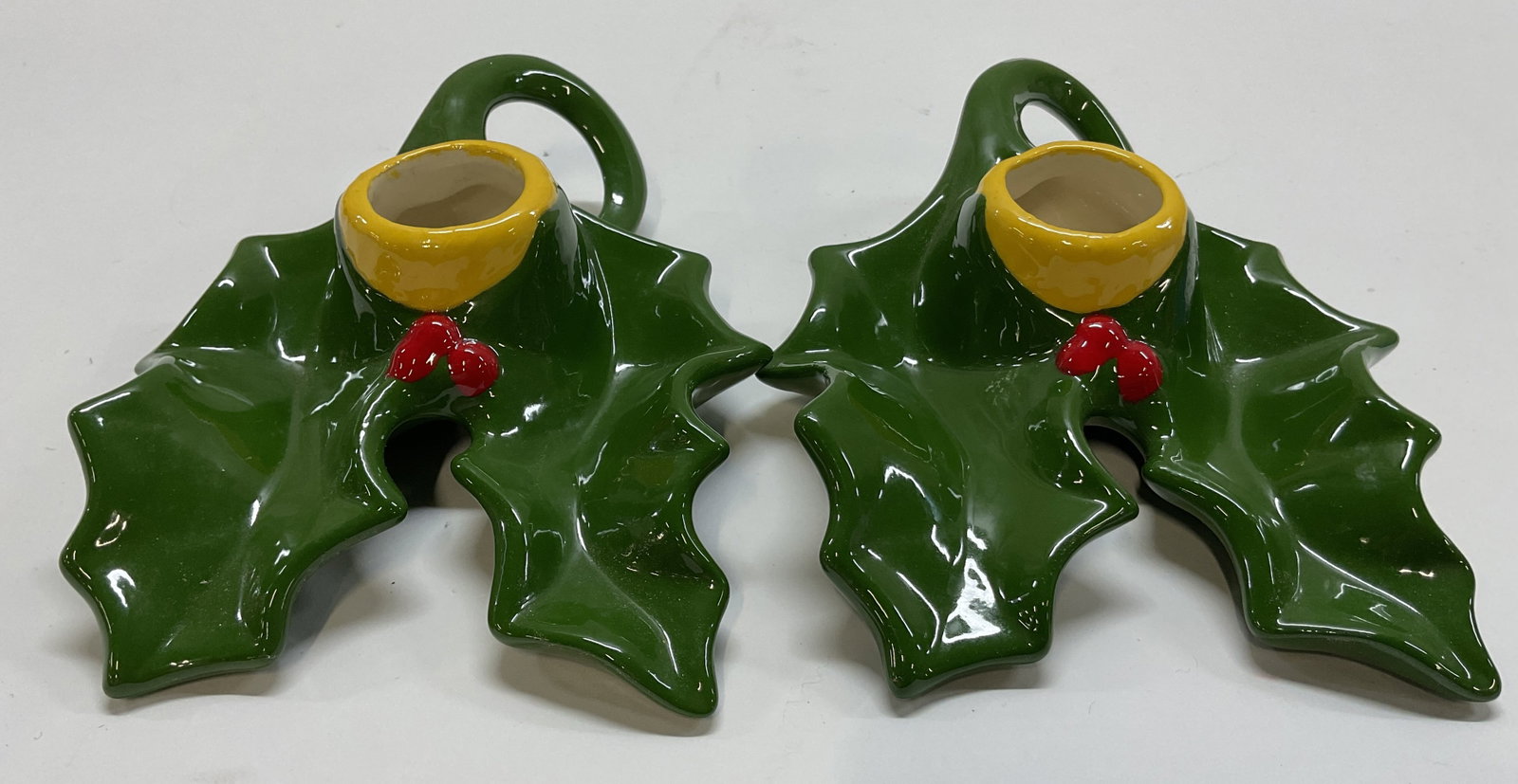 Pair Vtg Ceramic Xmas Holly Leaf Candle Holders: Pair of vintage ceramic Christmas holly leaf candle holders. Green glazed holly leaf shape with red berry accent. Yellow rimmed sockets. Each has an end handle. Scalloped edge and glossy finish.