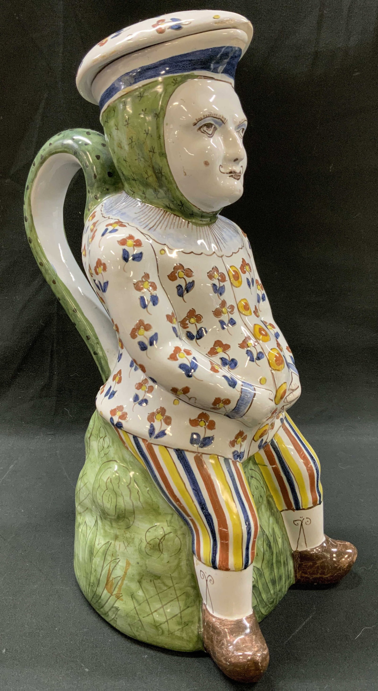 Vtg Majolica Lidded Character Toby Jug: Vintage hand painted majolica earthenware lidded character Toby jug. Modeled as a stylized man with a handlebar mustache, floral patterned jacket, striped trousers, sitting atop green grassy mound.