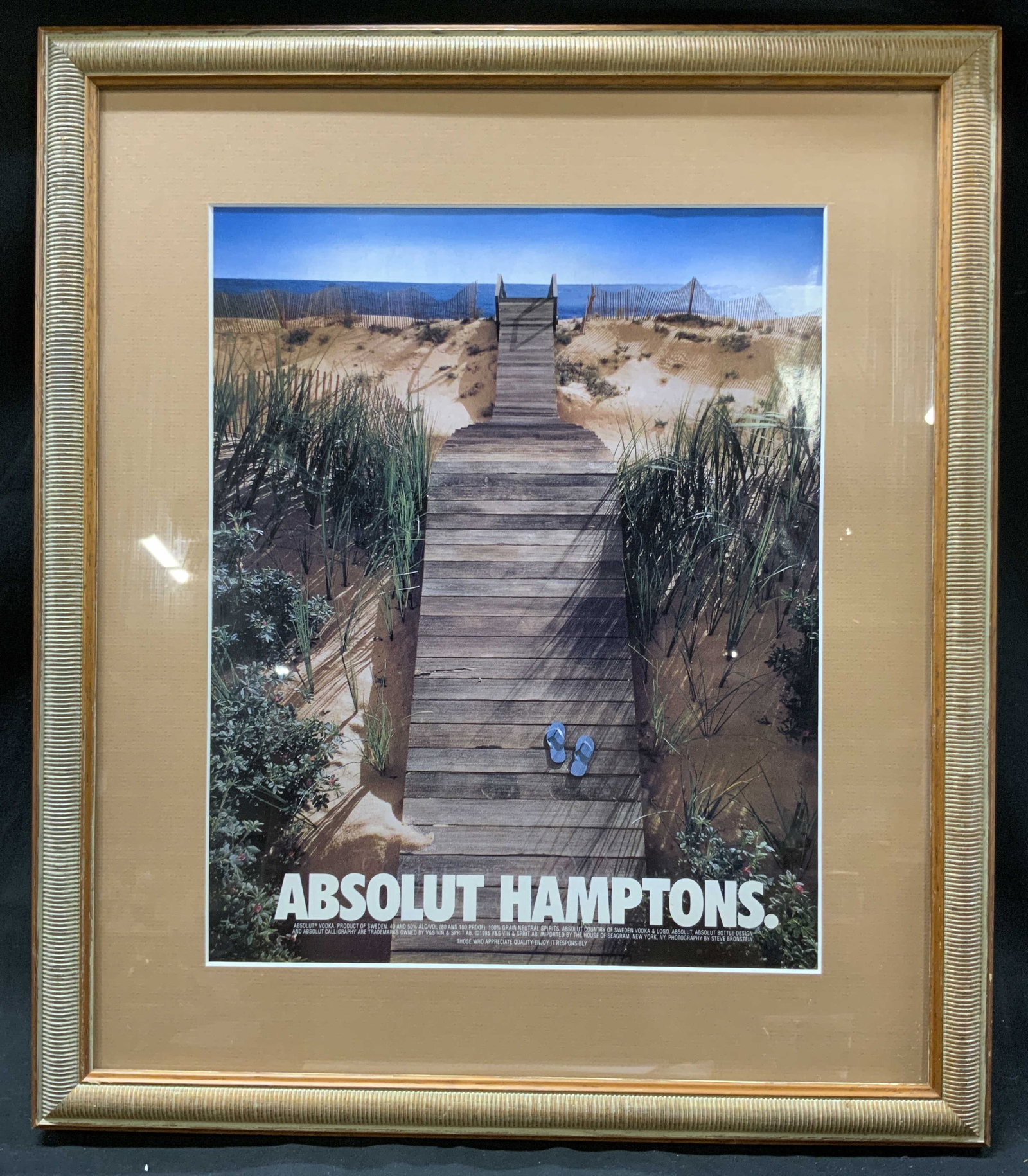 Vtg Frmd 1990s Absolut Hamptons Print Ad: Vintage late 1990s publication Absolut Hamptons campaign. Likely Sourced from a magazine. Wooden beach boardwalk, grassy dunes, pair of blue sandals. Boardwalk shaped into silhouette of Absolut Vodka