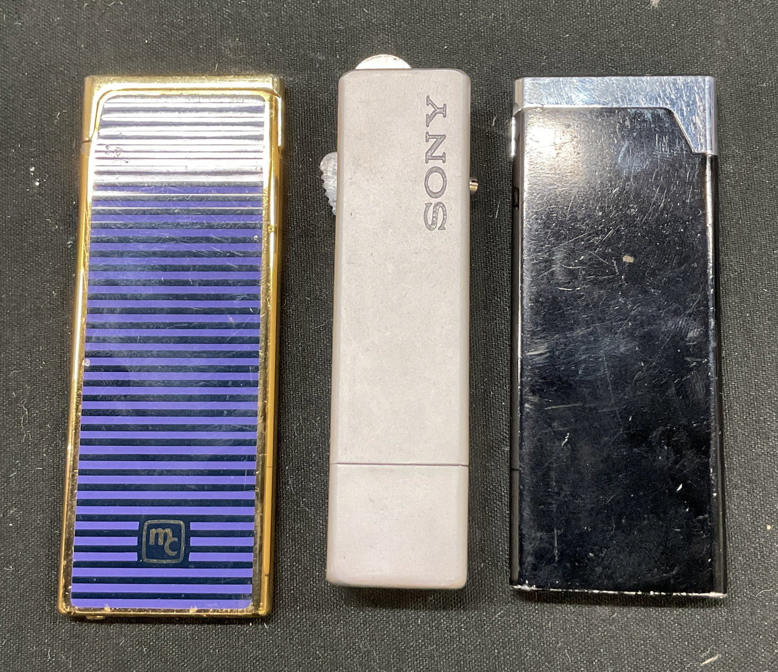 Lot 3 Vintage Sony Leo Limely, MC Lighters Japan: Lot of 3 vintage lighters. Brands feature Sony Leo Limely (Japan) and MC, marked on surface. Rectangular shaped. Silver toned, black, blue striped, gold toned. Measures approx 1.5 x 3 inches. Not test