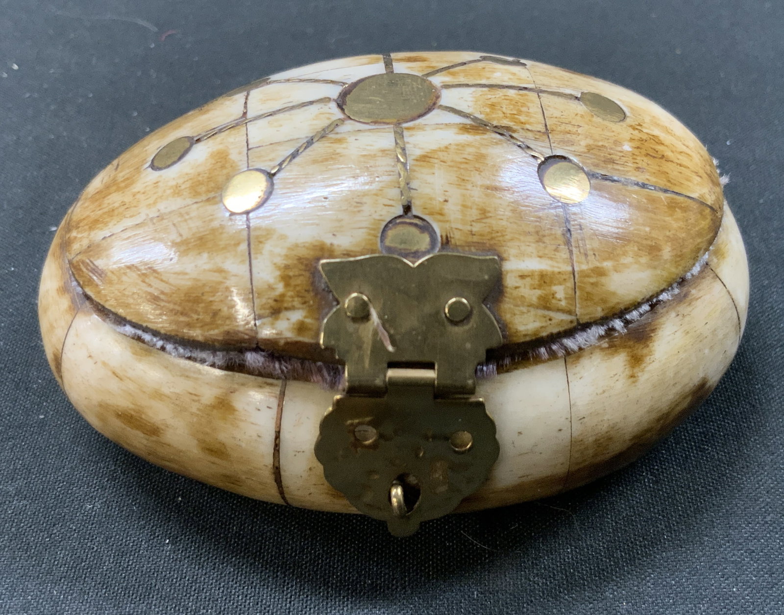 Vtg Bovine Bone Brass Inlaid Trinket Box: Vintage oval hinged pill/ trinket box. Multi panel bovine bone tile, domed lid with geometric starburst inlay of brass dots and lines. Opens on sturdy metal rear hinge. Interior purple velvet lined st