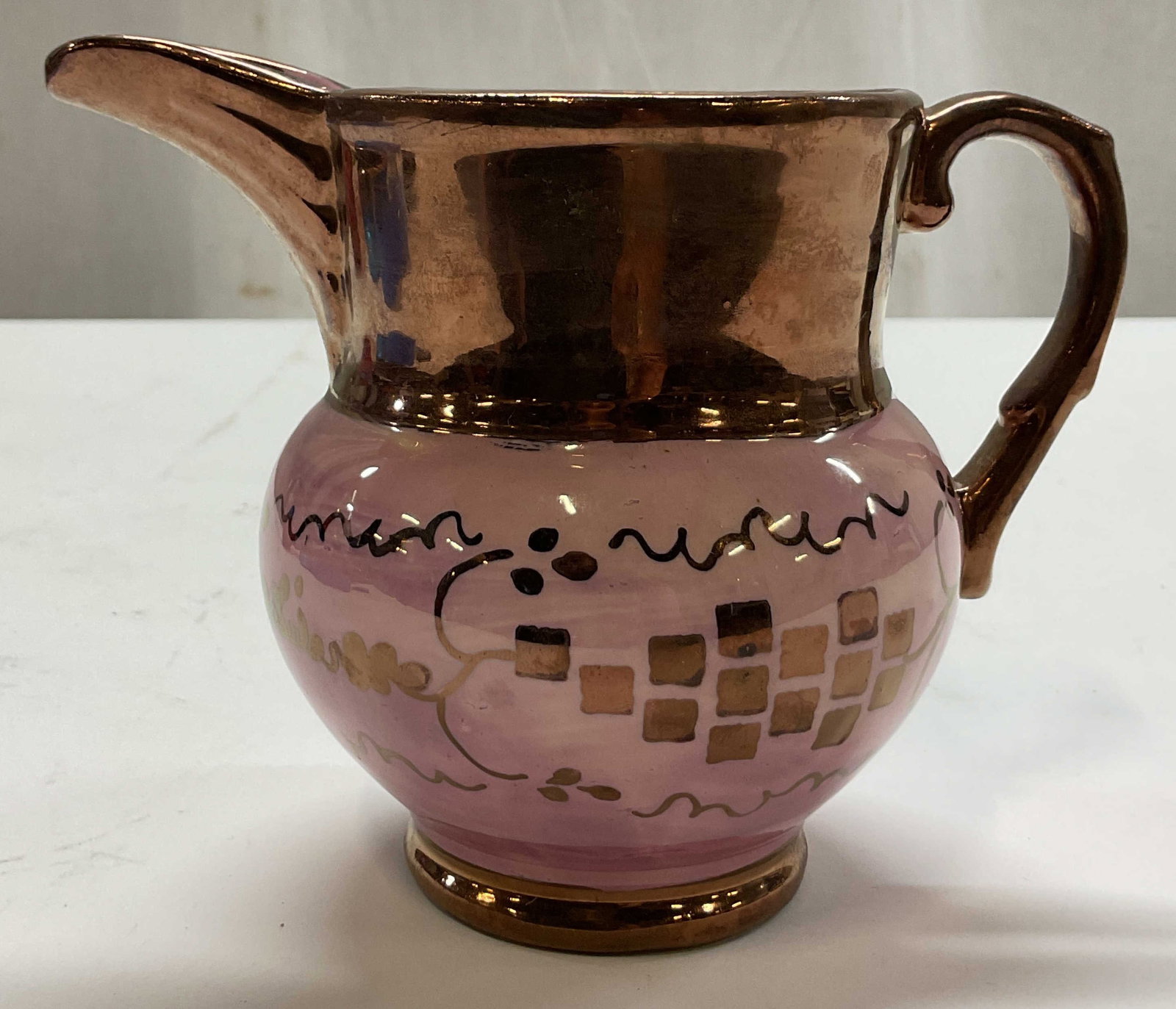 Cumbow Pottery Lusterware Pink Pitcher: Cumbow pottery Lusterware pink pitcher. Marked Cumbow on underside. Round base with curved handle and spout. Pink and purple with copper toned hand painted details and geometric patterns. Measures ht 