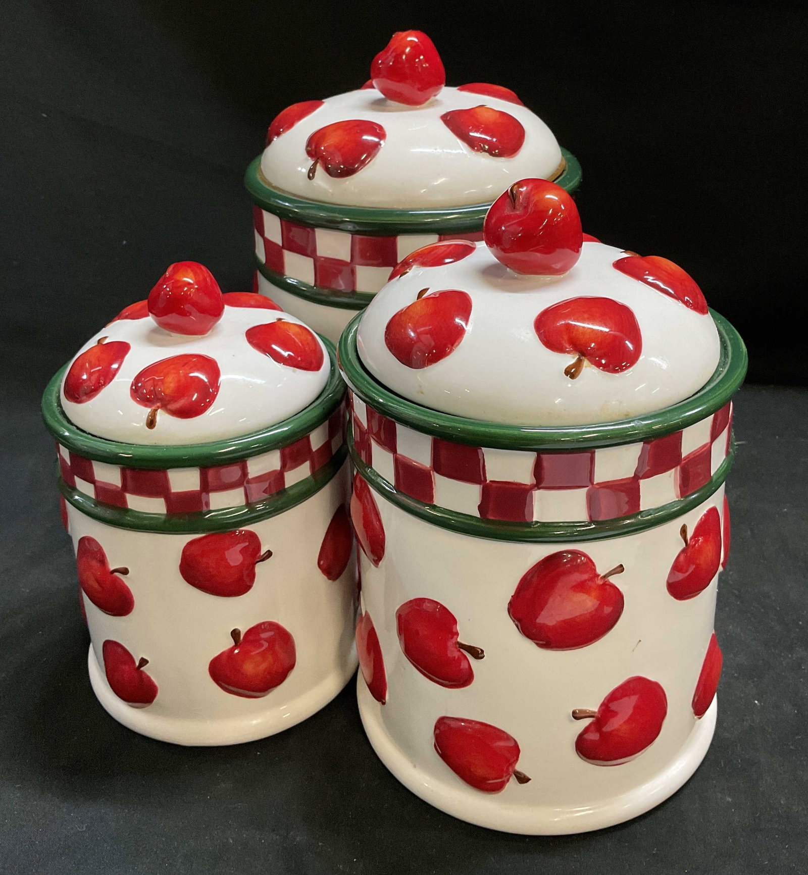 St3 Vtg New Debco Apple Canister Set: Set of 3 Vintage New Debco Ceramic Canister Set. Painted red apple patterns with red and green checkered rim. Apple knobs on lids. Some chips. 3 sizes. Largest measures app 8.5in H x 5.5in Diam.