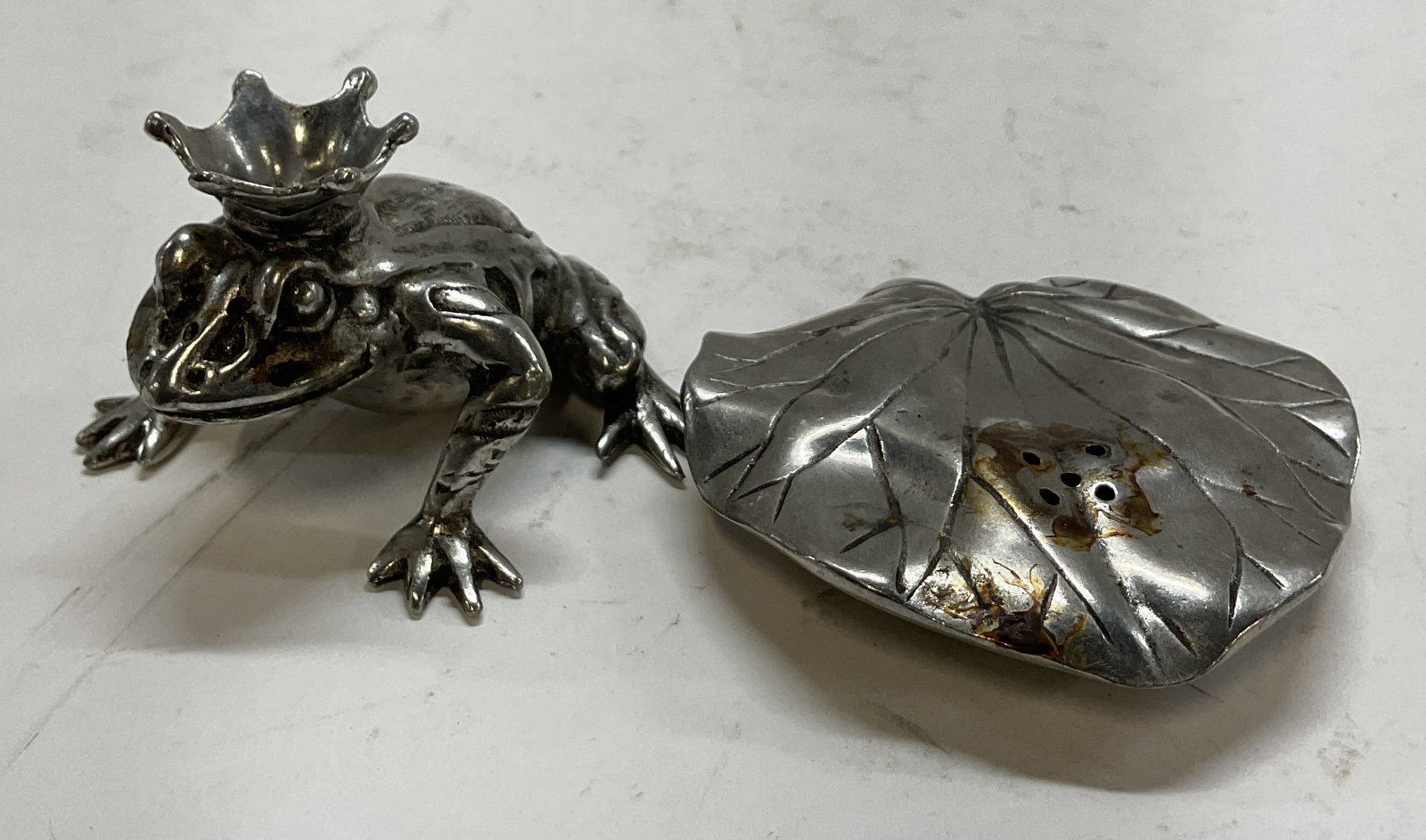 Vagabond House Frog Prince Salt & Pepper Shakers: Vagabond House frog prince salt and pepper shakers. Pewter material. Frog figure wearing a crown. Leaf figure. Detailed features and textures. Measures approx 3 x 3 inches. Scratching and surface