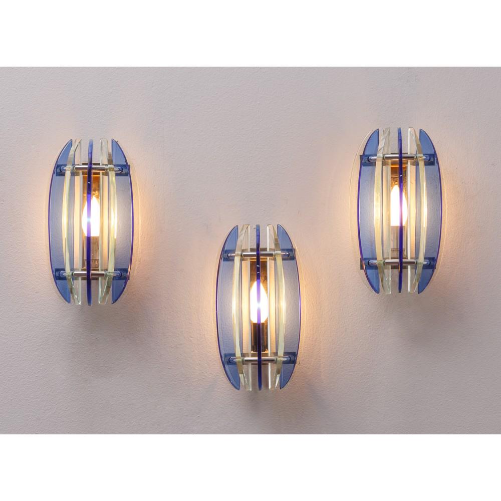 VECA, Tre lampade da parete: VECA Produzione Italia 1970 ca. Tre lampade da parete ad una luce, corpo ovalizzato in lame di vetro trasparente e blu struttura in metallo cromato, etichetta sul fondo. cm 16 x 10 x 27. THREE WALL LA