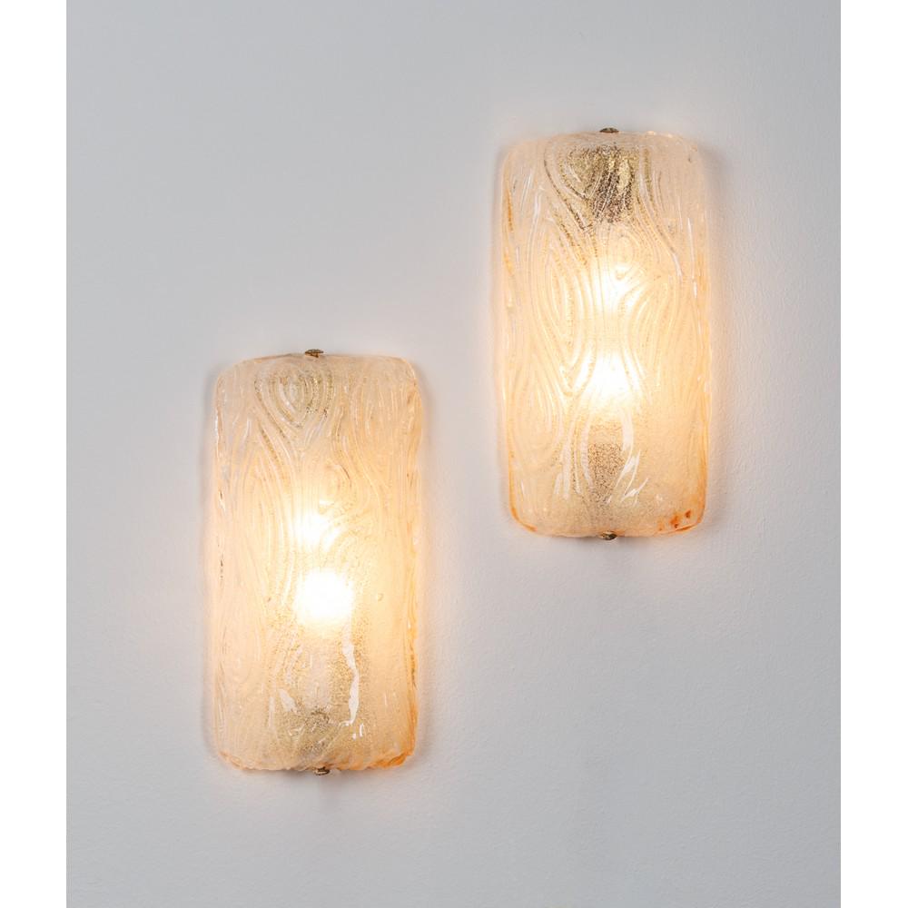 MAESTRI MURANESI, Coppia di lampade da parete: MAESTRI MURANESI Produzione Italia 1960 ca. Coppia di lampade da parete a due luci, corpo semicilindrico in vetro di grosso spessore decorato a rilievo con disegni geometrici astratti, supporto in met