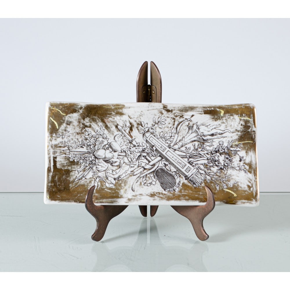 PIERO FORNASETTI, Svuota tasche in porcellana: PIERO FORNASETTI Produzione Italia 1950 ca. Svuota tasche in porcellana con decoro nero su fondo oro ispirato alla classicità. Usure. cm 25 x 12 Alt. 2,5.