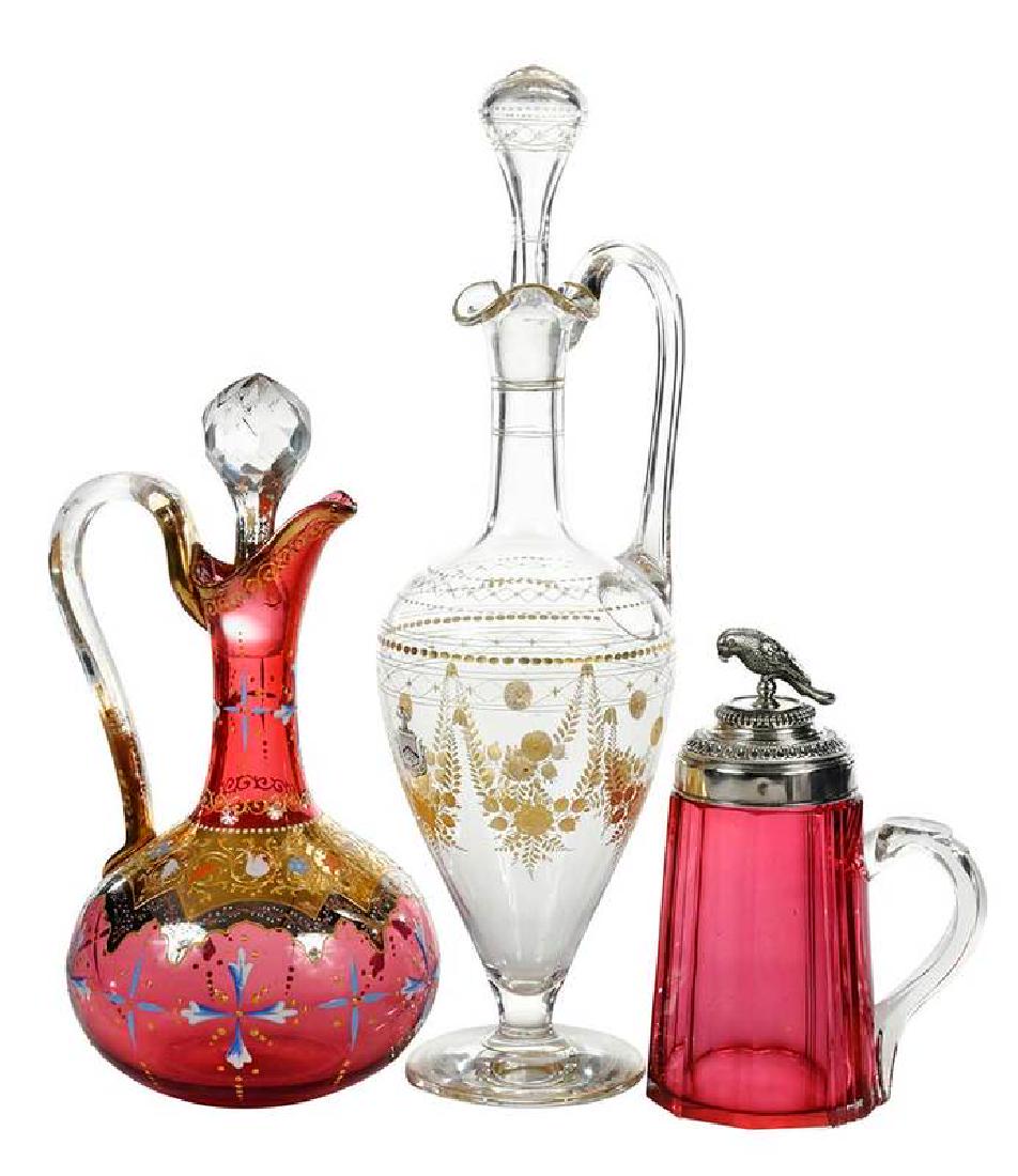 Three Decorated Glass Table Items: handled ewer with stopper, cut gilt floral swags and accents, small figure over shield and banner, "Pro Deo Et Patria" (For God and Country), 17 in.; Moser enamel and gilt decorated cranberry ewer wit