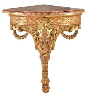 Italian Baroque Mask Carved Corner Console: probably Roman, 18th century, with quarter round variegated marble top over well carved gilt support centering a satyr mask flanked by bellflower garlands, above a single drapery and leaf carved leg,