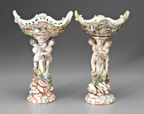 Two similar porcelain tazza: each with pedestal with two [putti] at tree trunk on rocky ground below reticulated basket with applied flowers, one Meissen with blue crossed swords mark under glaze with two hash marks below (possib