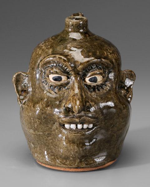 Lanier Meaders face jug: (White County, Georgia, 1917-1998), alkaline glaze stoneware, mottled olive glossy glaze, ceramic teeth, painted eyes, detailed eyebrows, marked in script on base "Lanier Meaders", 9-3/8 in. Excellent