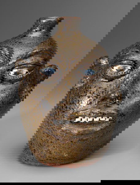 Lanier Meaders Face Jug: Georgia, 20th century, stoneware, mottled olive-green glaze, ceramic teeth and eyes, incised eyelash and eyelid detail with large pronounced eyebrows, large lips, pronounced nose with large nostrils,