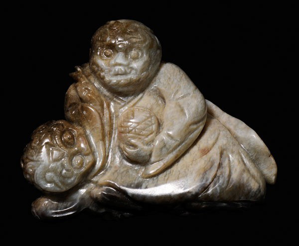 Jade [Lohan]: Qing Dynasty, partially burnt grayish-beige "chicken bone" jade, triangular-shaped with olive and brown areas, seated [lohan] with Buddhist lion behind him looking around his right side at a brocade b