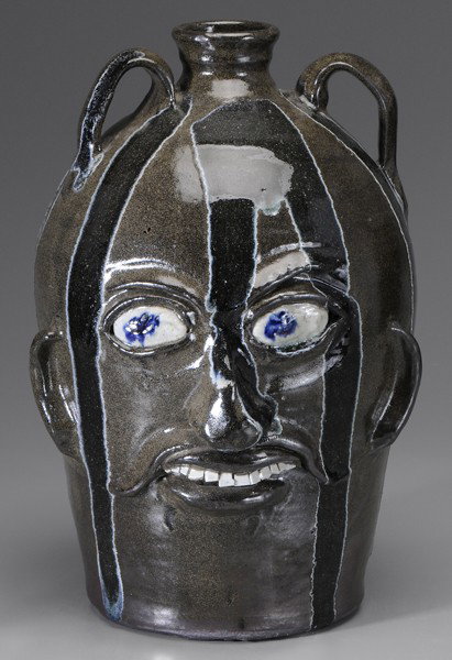 Chester Hewell Face Jug: (Gillsville, Georgia, born 1950), dark and light glaze runs over medium olive glaze, two strap handles, ears, blue-painted eyes, broken-china teeth, marked on bottom in script "Chester Hewell CSA 1977