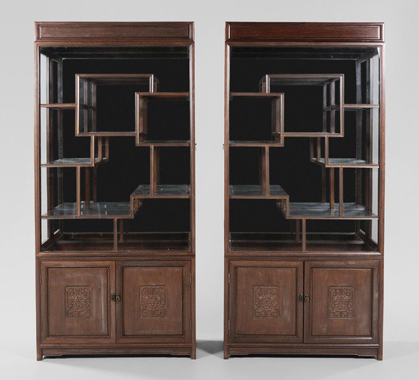 Pair Ming Style Carved Mahogany: Cabinets, Chinese, 20th century: each with lighted removable cornice over a mirror-backed cabinet with side doors and glass shelves, over pedestal base with two carved and paneled doors and shelved in