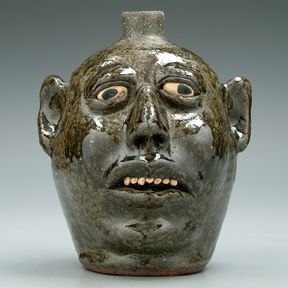 Lanier Meaders stoneware face jug,: painted bulging eyes, ceramic teeth, runny medium olive alkaline glaze, base marked in script "Lanier Meaders", 8-1/2 x 7 in. Some glaze irregularities.